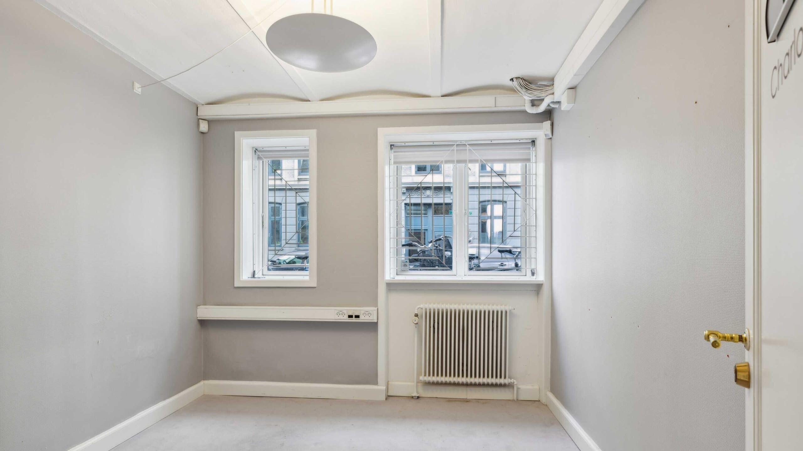 Et tomt rum med to vinduer og en radiator. På gulvet er der en mørk gulvbelægning. Væggene er hvide, og der er et rundt