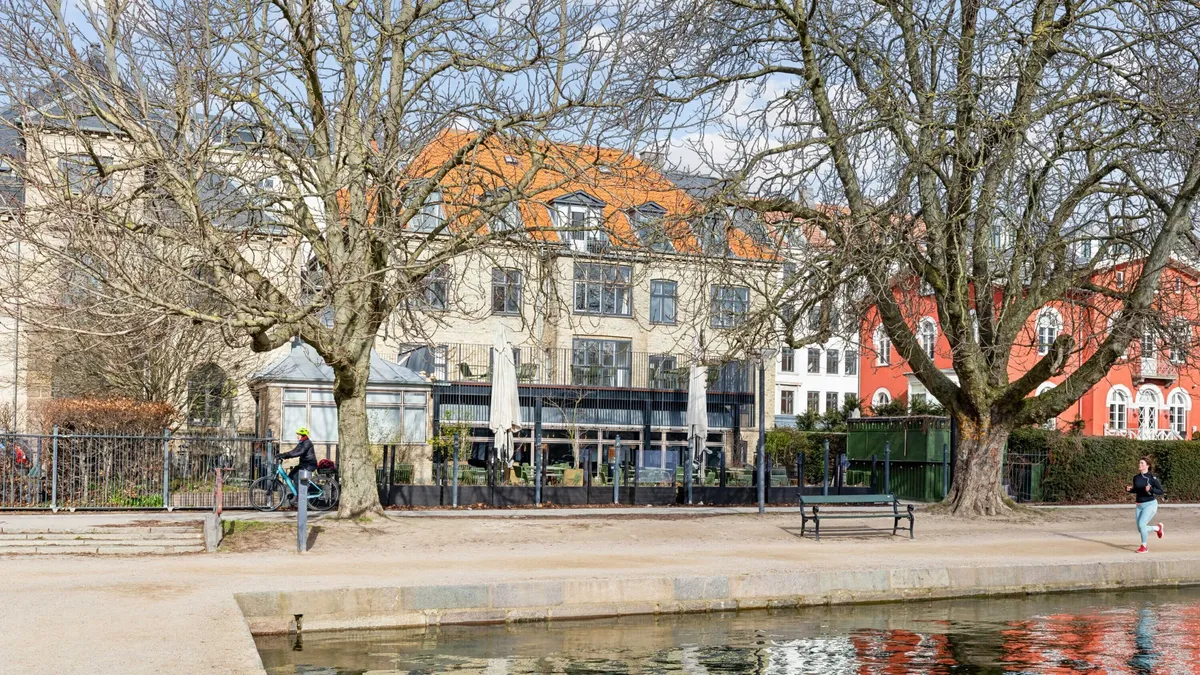 Et søsideområde med en gående person og en person på cykel langs vandkanten, og flere bygninger i baggrunden