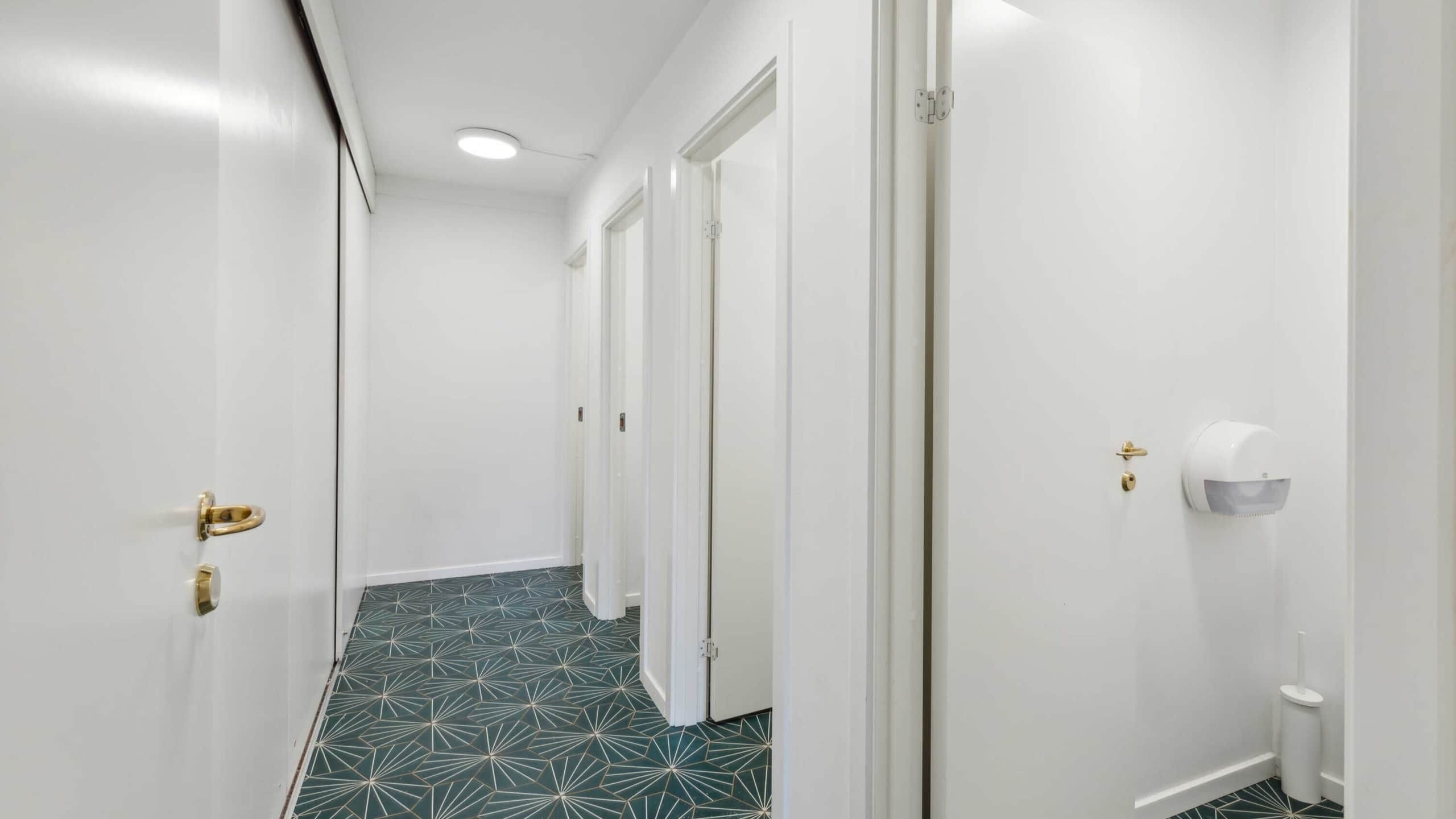 Langt og smalt korridor med hvide vægge og gulv med grønt og guld mønster samt tre lukkede hvide døre