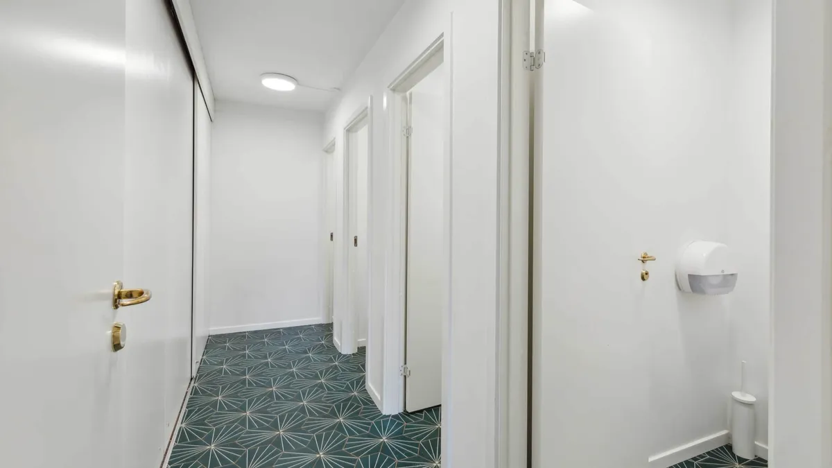 Langt og smalt korridor med hvide vægge og gulv med grønt og guld mønster samt tre lukkede hvide døre