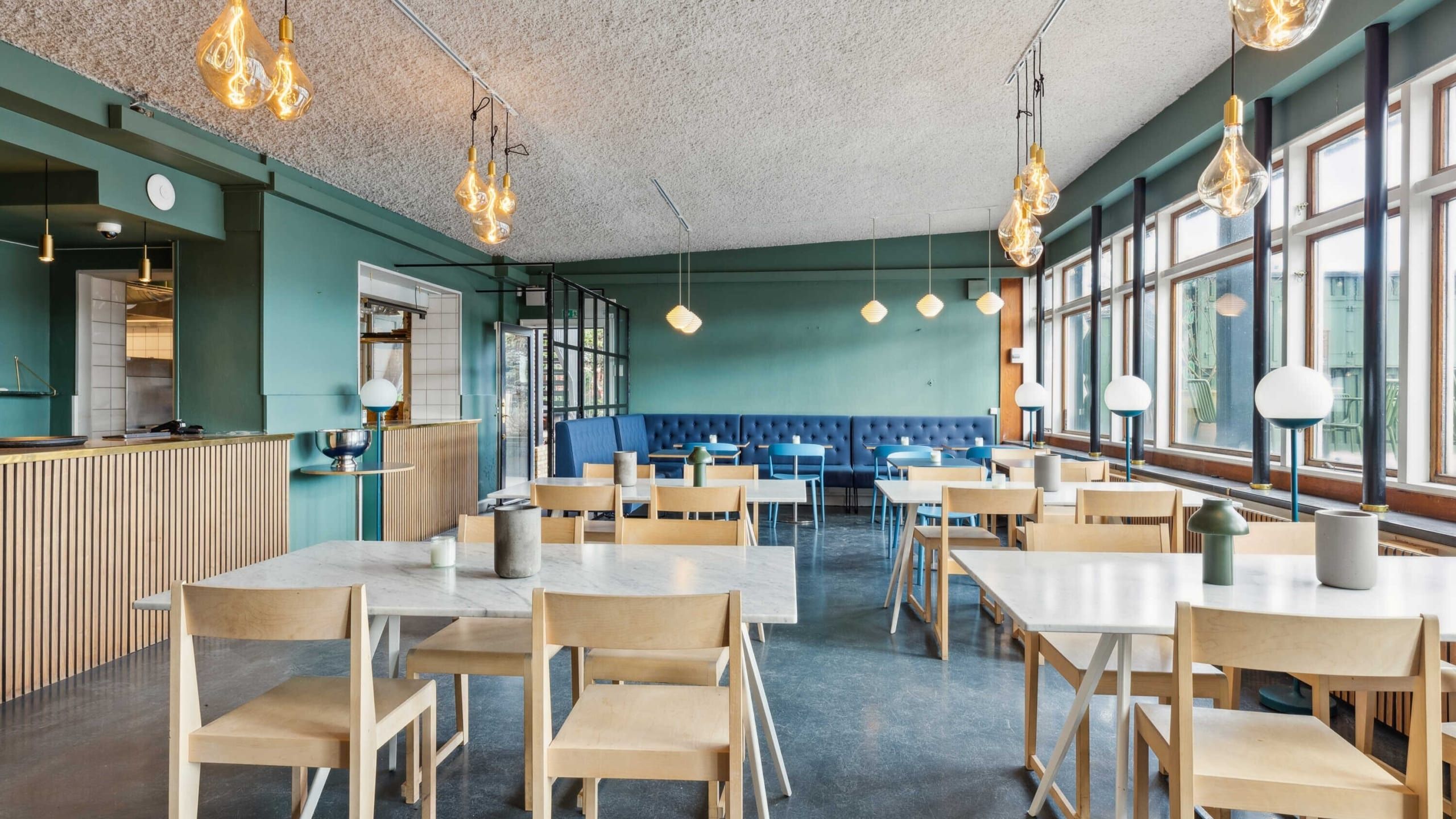 Interiør med lysegrå vægge og hvide borde og stole, pendellampe med lyse pærer og lysekroner og vinduer med udsigt til gaden