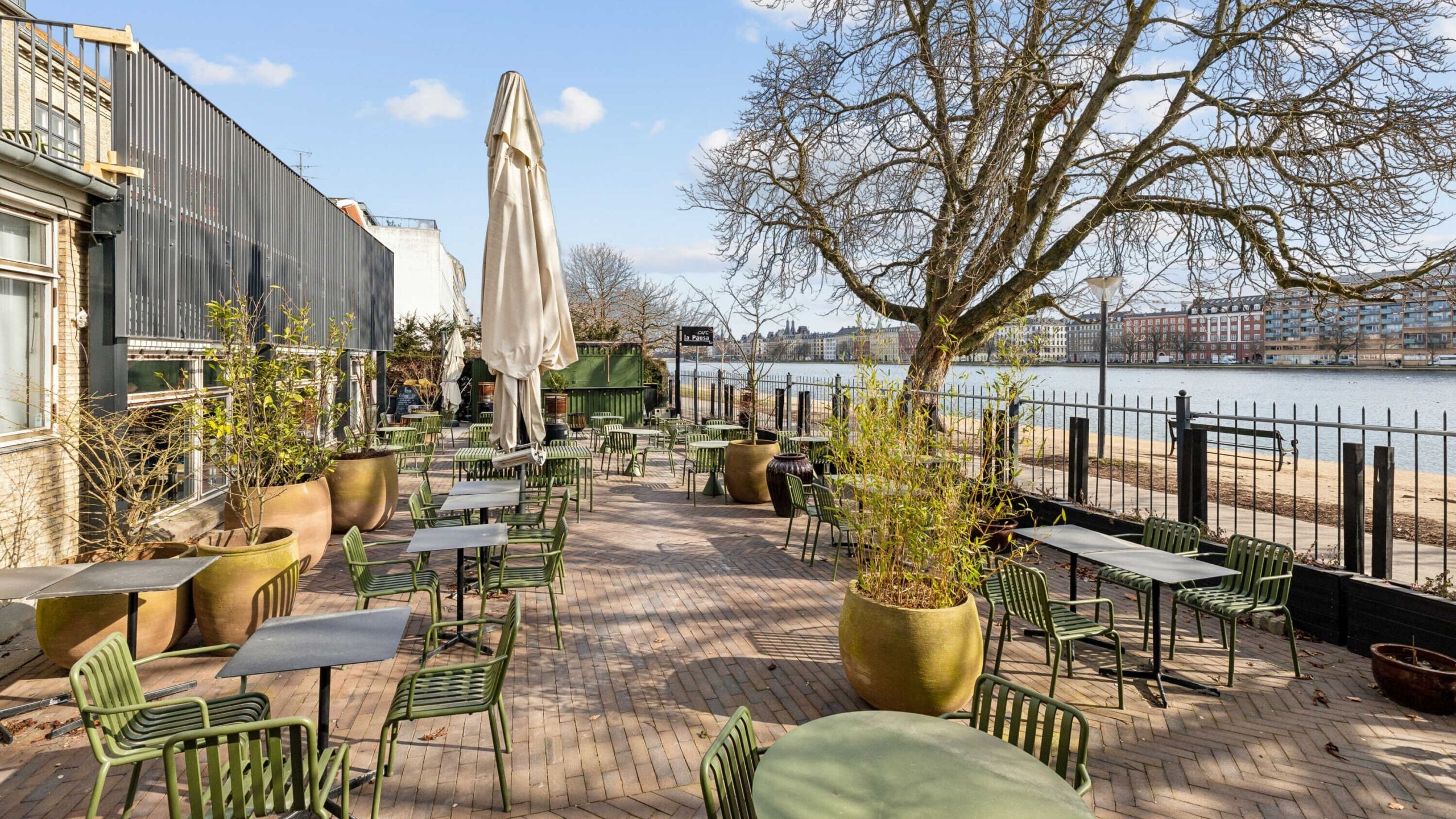 En kafé med grønne stole og borde placeret ved en flod med et hav af bygninger i baggrunden