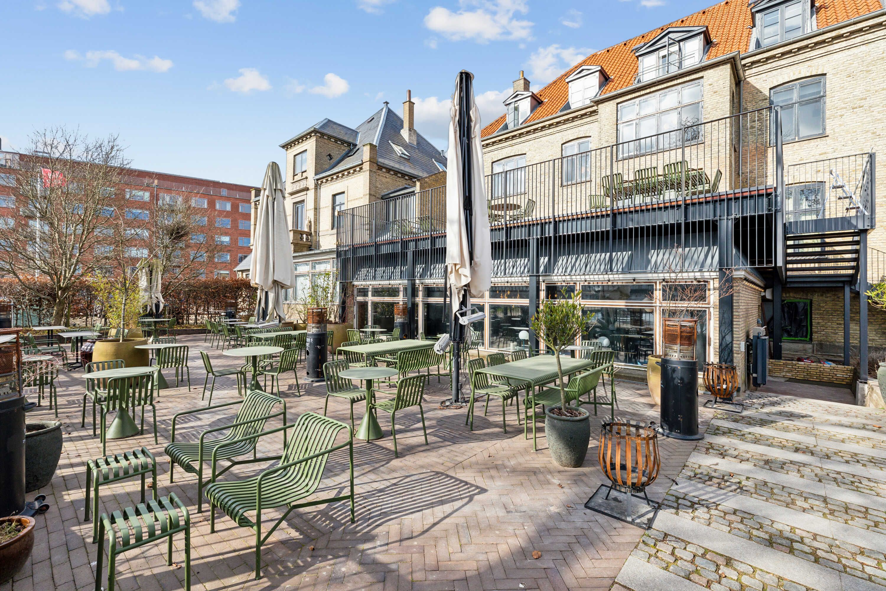 Terrasse med grønne stole og borde, lukket paraply og plantepot, lyserødt tag på bygning og lyserøde taglejer