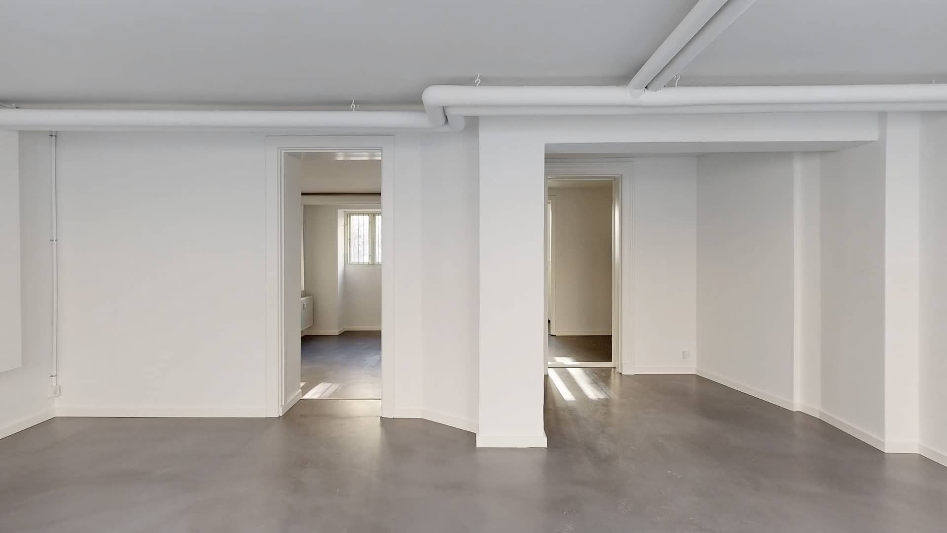 Store, tomt værelse med hvide vægge og gulv, højt loft med hvide rør og lysestråler fra vinduet