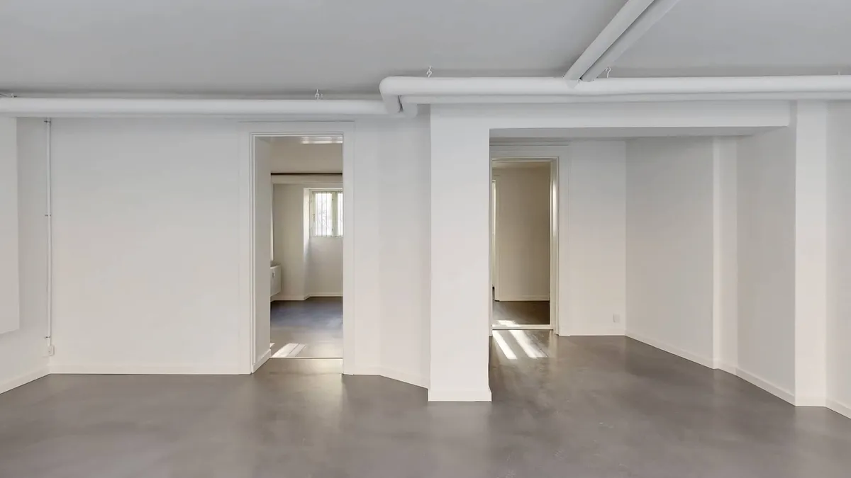 Store, tomt værelse med hvide vægge og gulv, højt loft med hvide rør og lysestråler fra vinduet