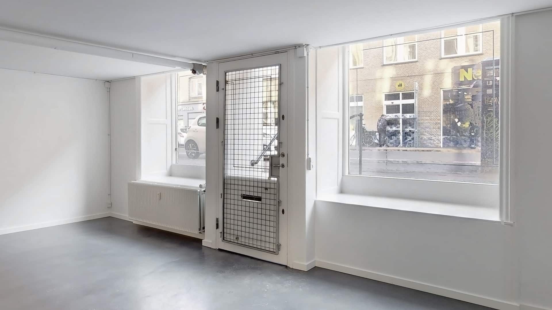 Et tomt lokale med hvide vægge og gulv, store ruder og en dobbeltdør i metalgitter. En radiator er monteret på væggen ved