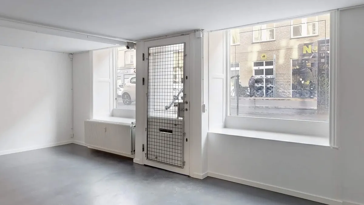 Et tomt lokale med hvide vægge og gulv, store ruder og en dobbeltdør i metalgitter. En radiator er monteret på væggen ved