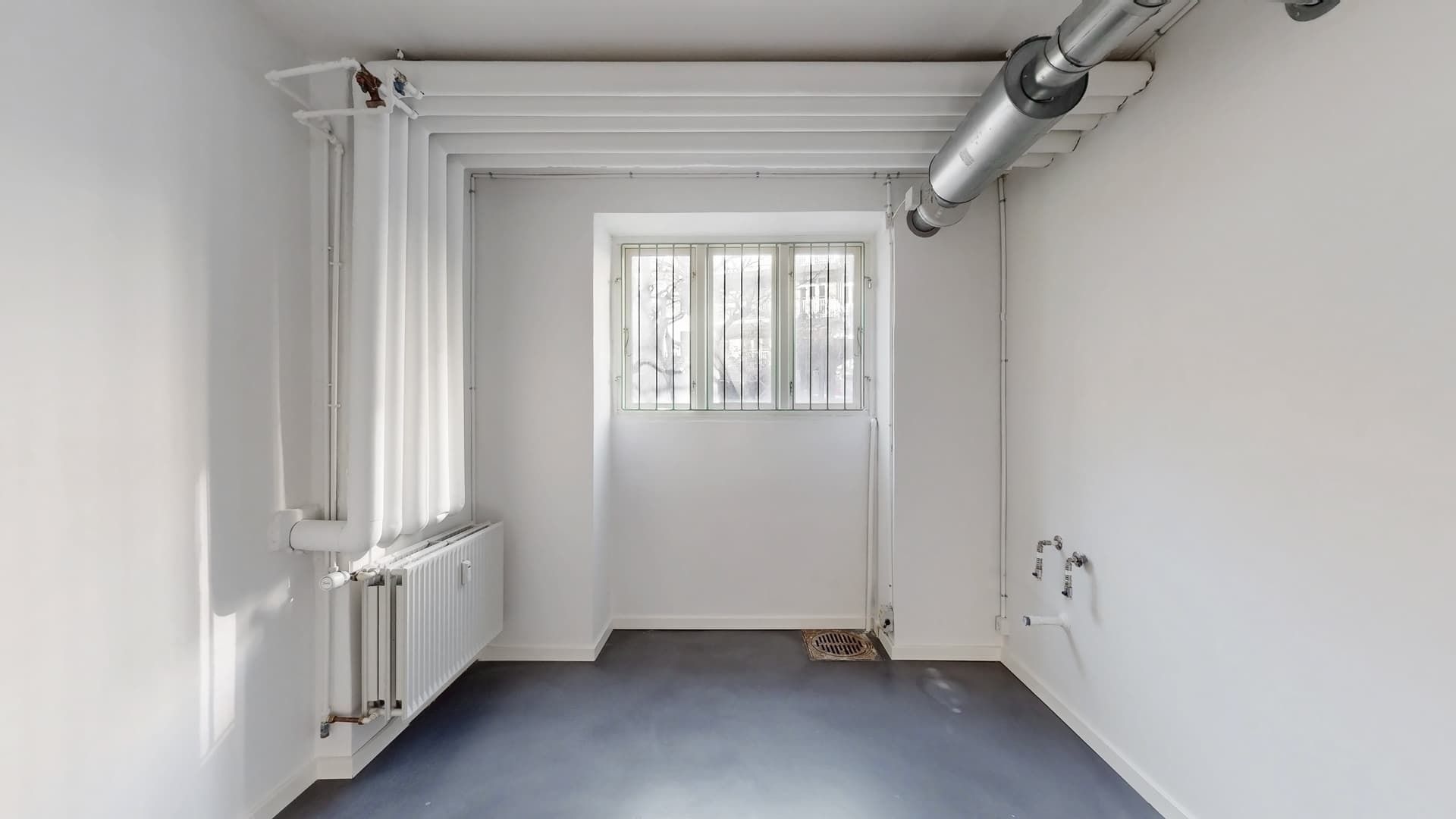 Et tomt rum med hvide vægge, sort gulv og et vindue med gitter. Et radiator er monteret på den ene væg og et ventilrør er