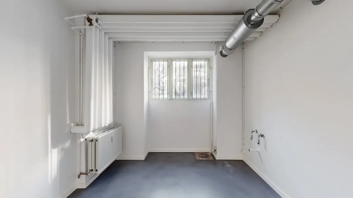 Et tomt rum med hvide vægge, sort gulv og et vindue med gitter. Et radiator er monteret på den ene væg og et ventilrør er