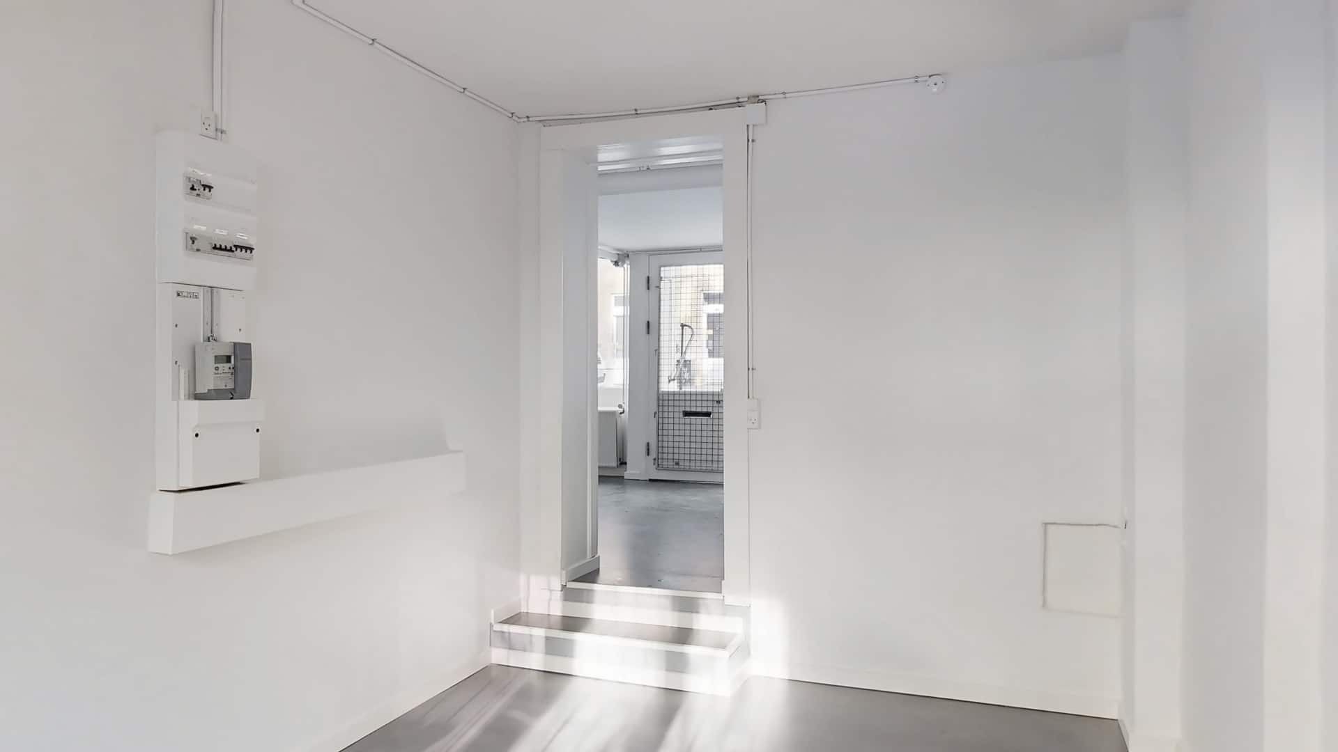 Indvendigt erhvervslejemål med hvide vægge og gulv samt åben dør til et værelse med trin.