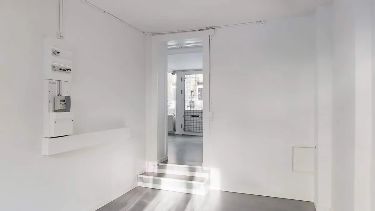 Indvendigt erhvervslejemål med hvide vægge og gulv samt åben dør til et værelse med trin.