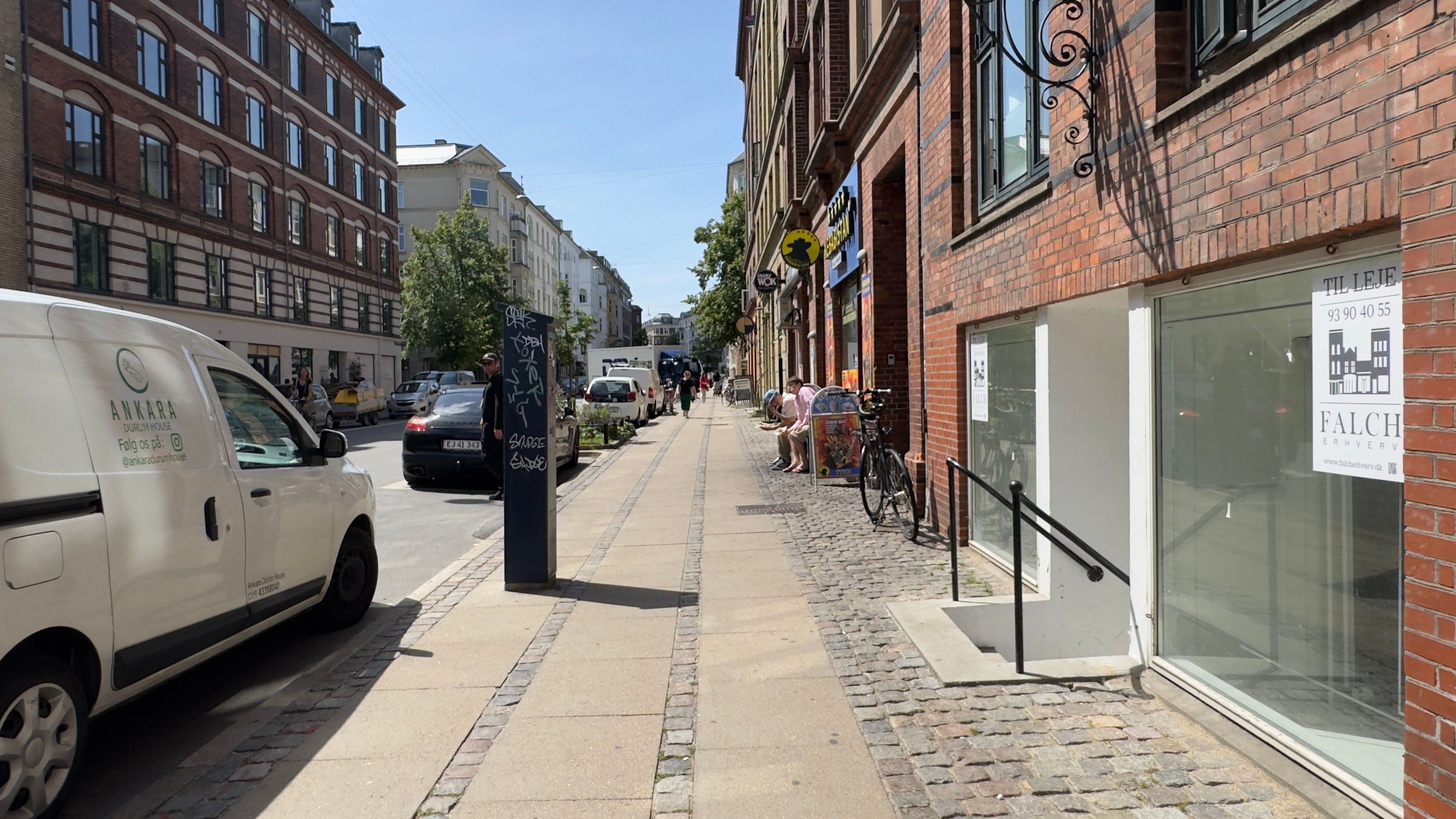 Bred sidegade med høje bygninger, forretninger og parkerede biler, folk går, og to mennesker sidder på en stol udenfor en