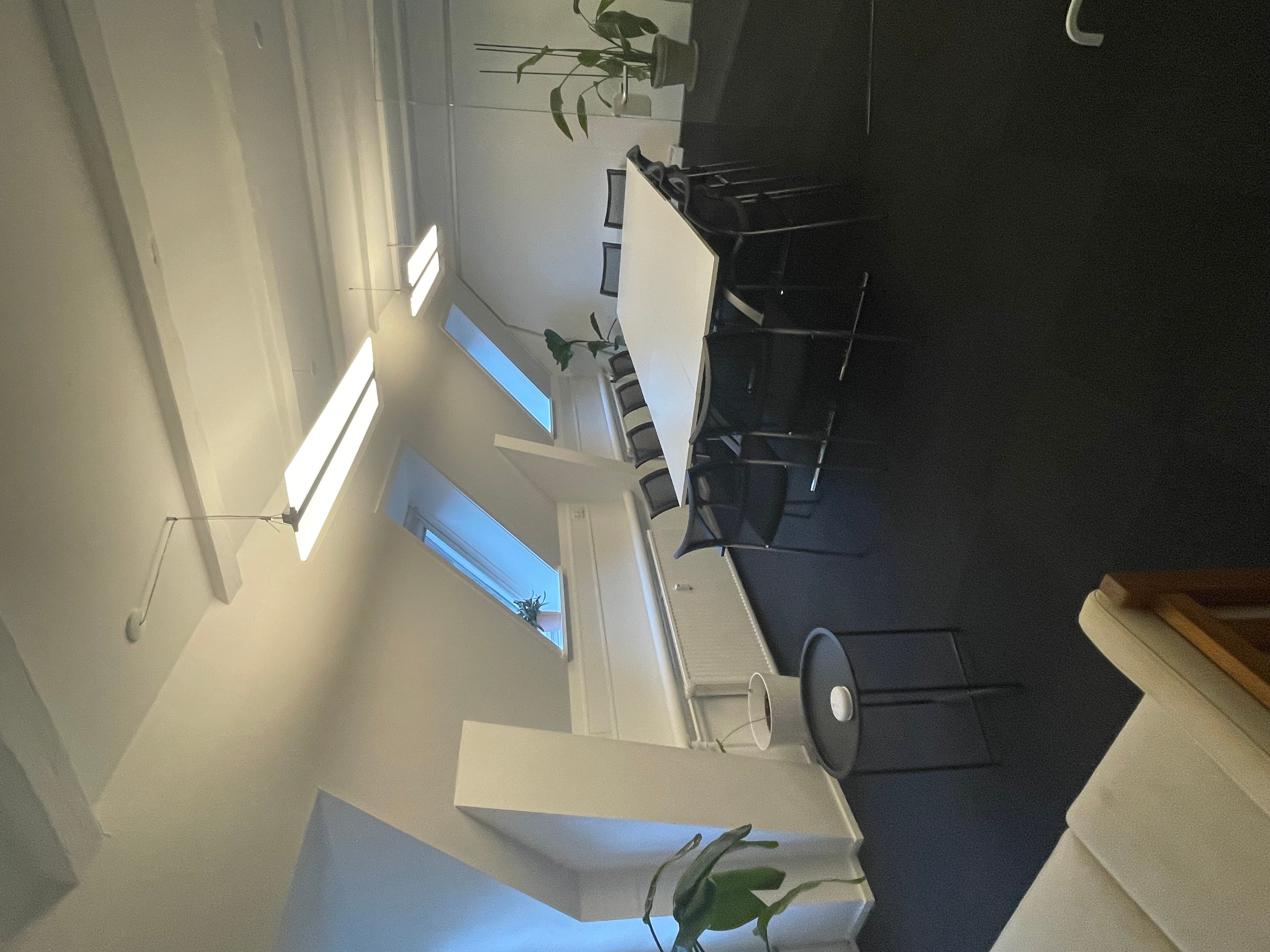 Et kontor med et stort hvidt bord og flere stole samt plante i hjørnet og lysekroner på loftet