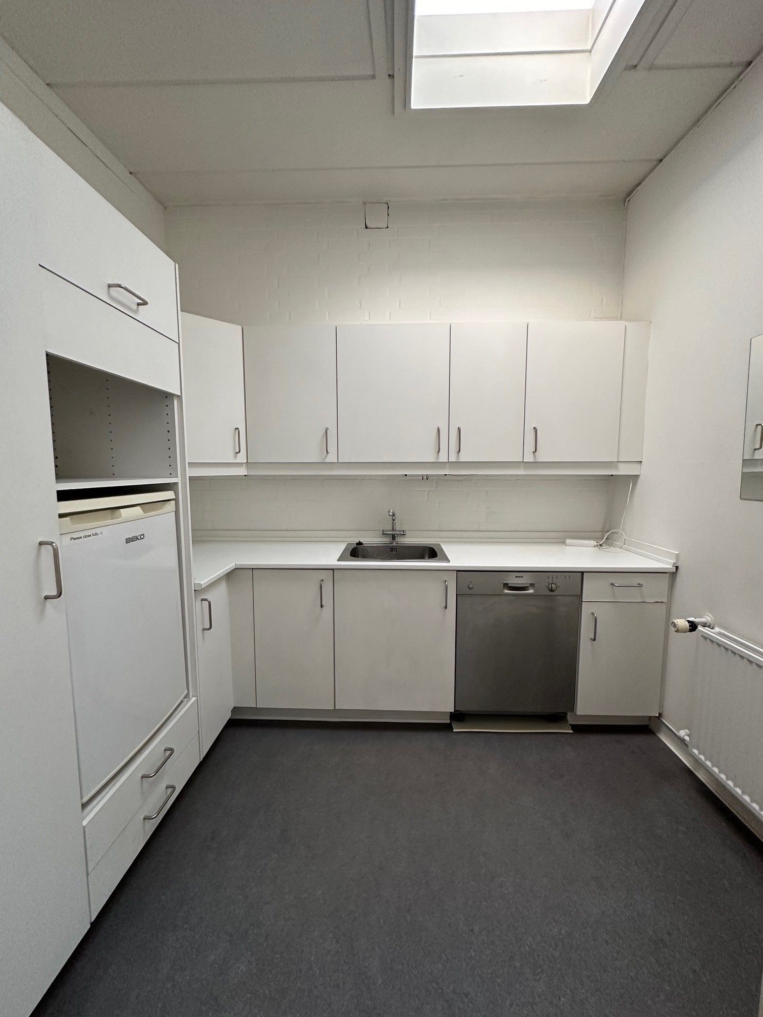 Moderne køkken med hvide skabe, brædder og højloft med loftsvindue, ren overflade med vandhaner, vaskemaskine, køleskab og