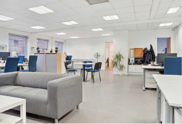 Kontorlokaler med lyse møbler, højloft og glasvinduer, hvor flere personer arbejder på computere og skriveborde