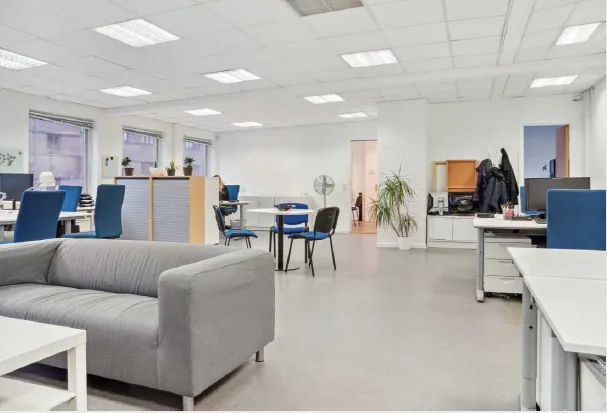 Kontorlokaler med lyse møbler, højloft og glasvinduer, hvor flere personer arbejder på computere og skriveborde