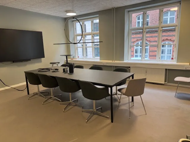 Mødelokarium med skrivebord, stole og en stor tv-skærm på væggen. Der er vinduer med udsigt til bygninger og radiatorer