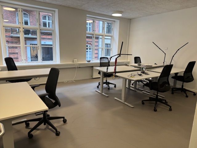 Et kontor med hvide vægge og gulv. Bord, stol og lampe er placeret. Et vindue giver udsigt til en bygning med en række