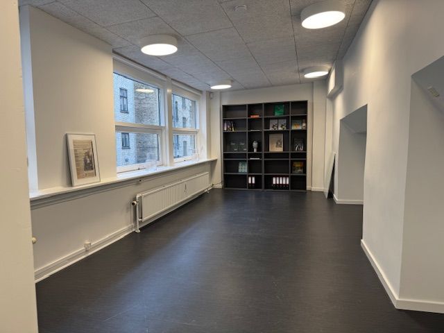Et tomt kontor med hvide vægge og gulv samt vinduer med udsigt til en bygning. Der er et stående kabinet med bøger og en