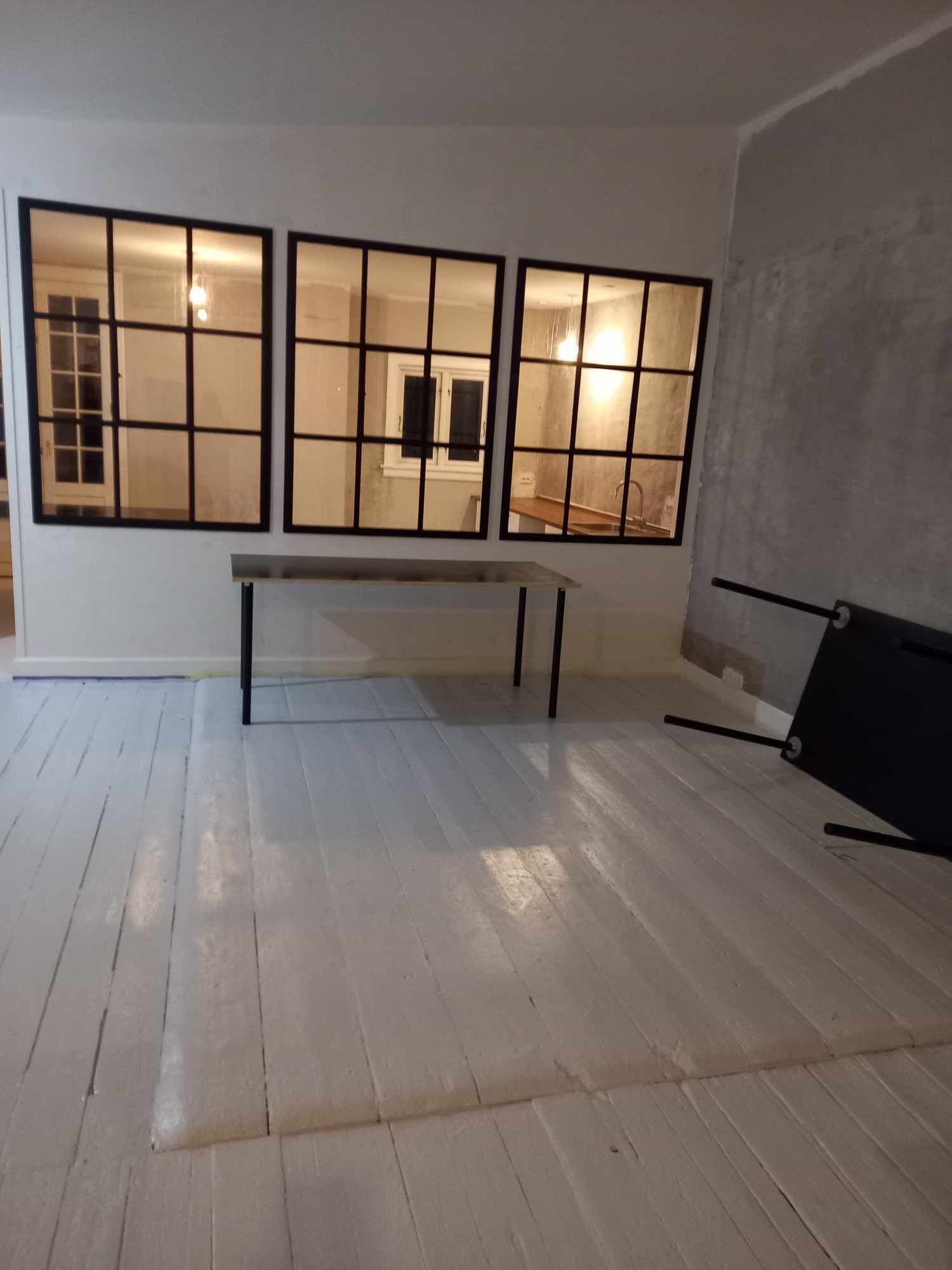 Et rum med lyse gulvbelægning og lyse vindue med træindfatning og sort kant. Der er en stor, simpel bord og et faldet stol