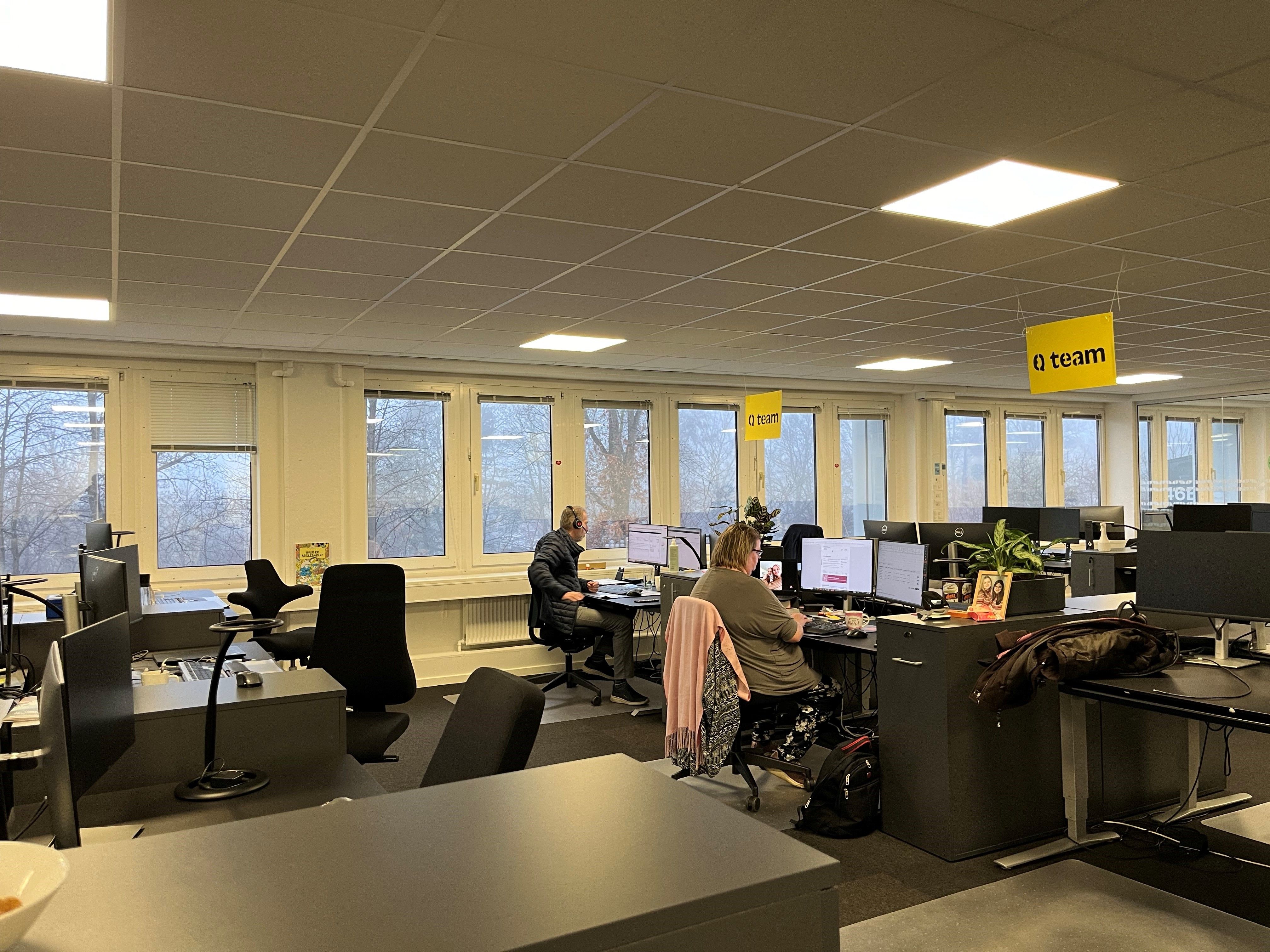 To personer sidder og arbejder i en moderne kontorforretning med solbeskyttelsesrullegardiner og lyse loftslamper.