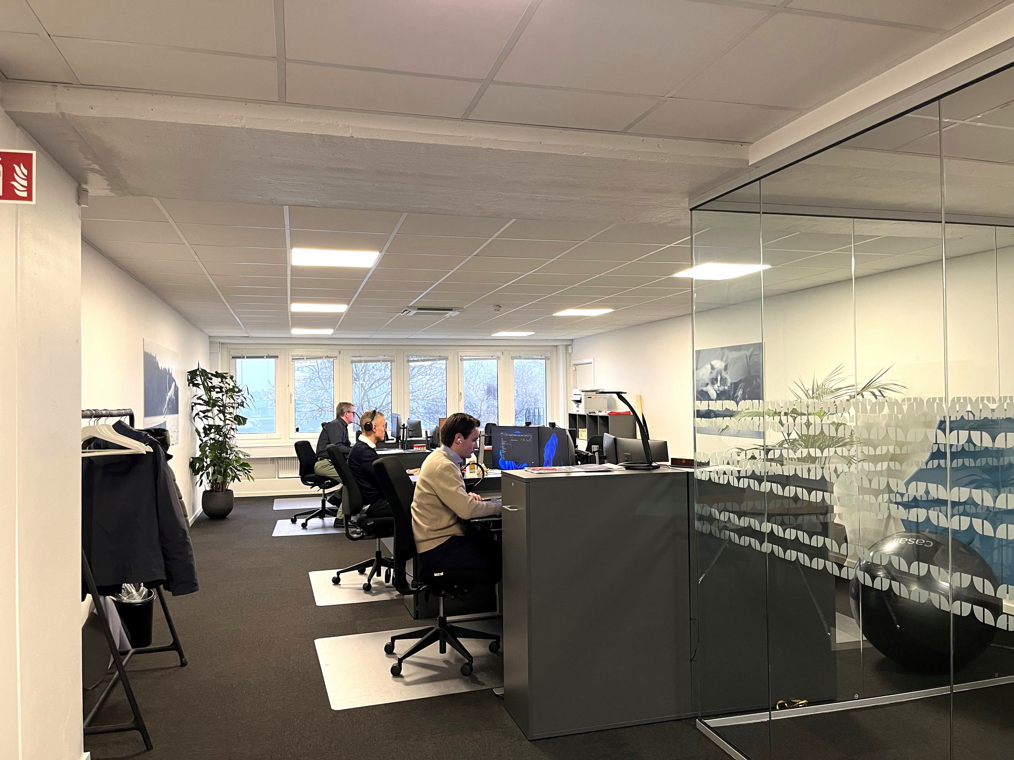 Kontorrum med moderne møbler og fire personer, der sidder og arbejder ved computere, med en stor glasvæg og indvendige