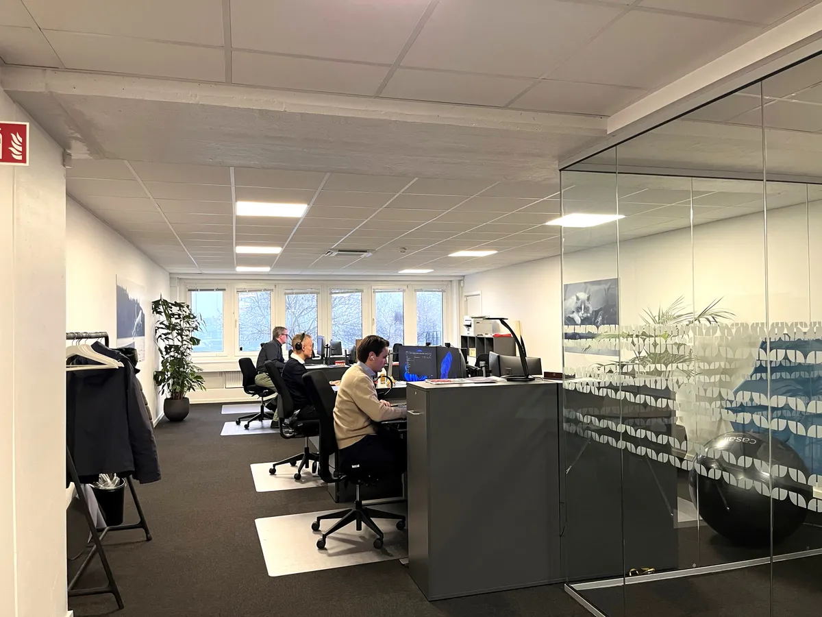 Kontorrum med moderne møbler og fire personer, der sidder og arbejder ved computere, med en stor glasvæg og indvendige