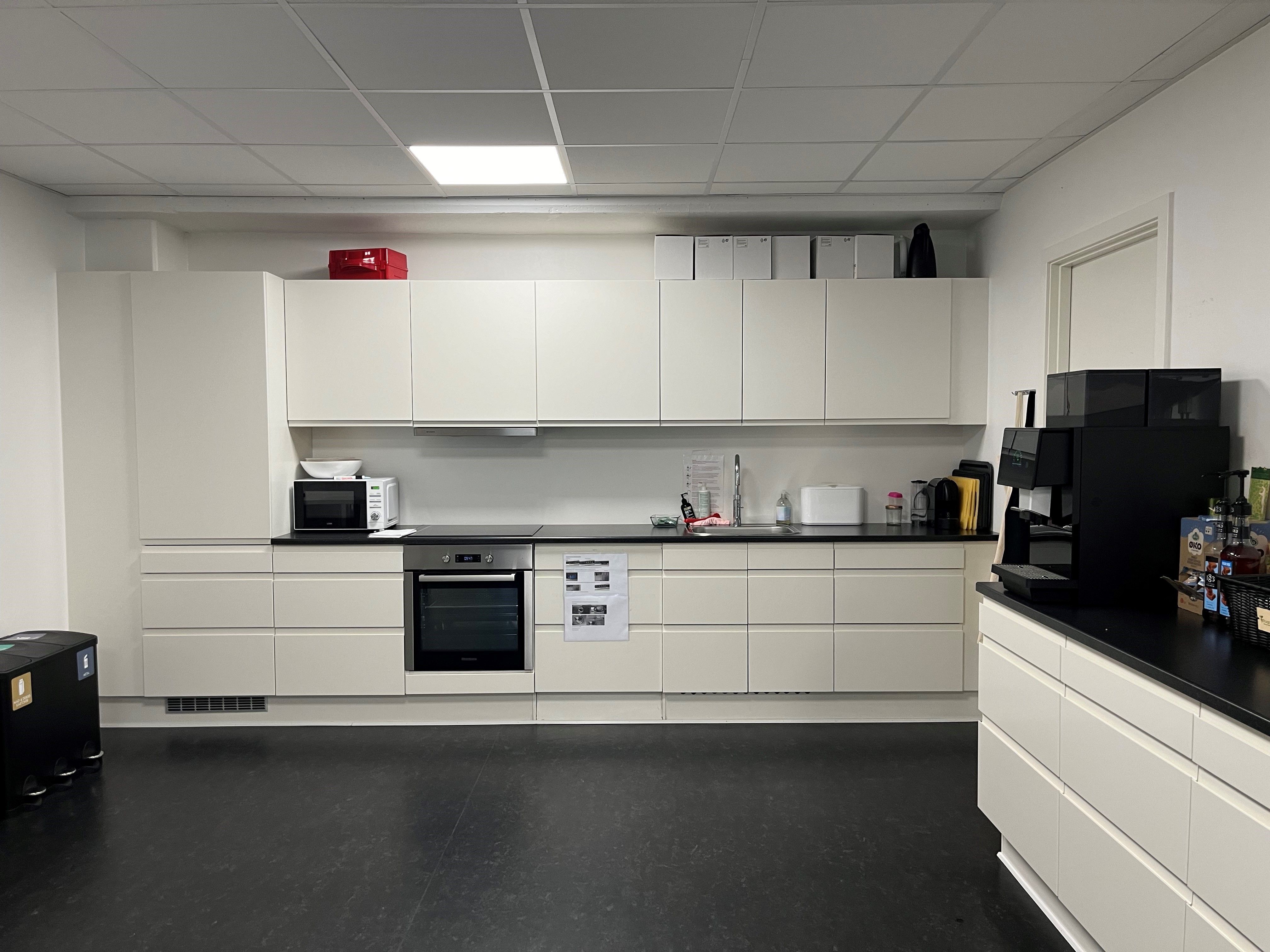 Kontorlejemål med moderne køkken og højloft, hvidt køkken med indbygget ovn og mikroovn, sort kaffemaskine, alle hængende