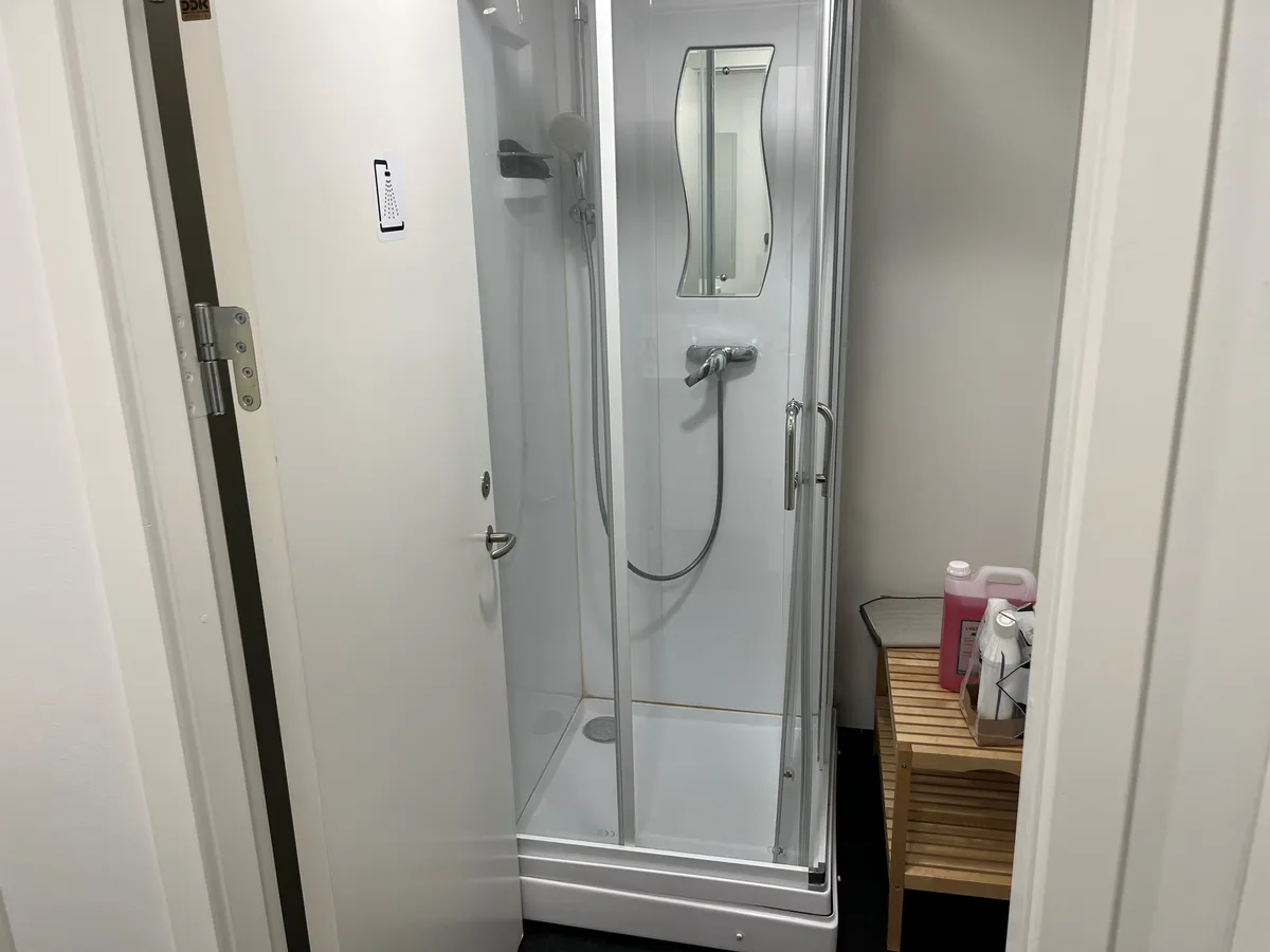 En hvid enkle og moderne brusekabine med en indbygget spejl, hånddusch og håndledning samt et træbord med rengøringsmidler