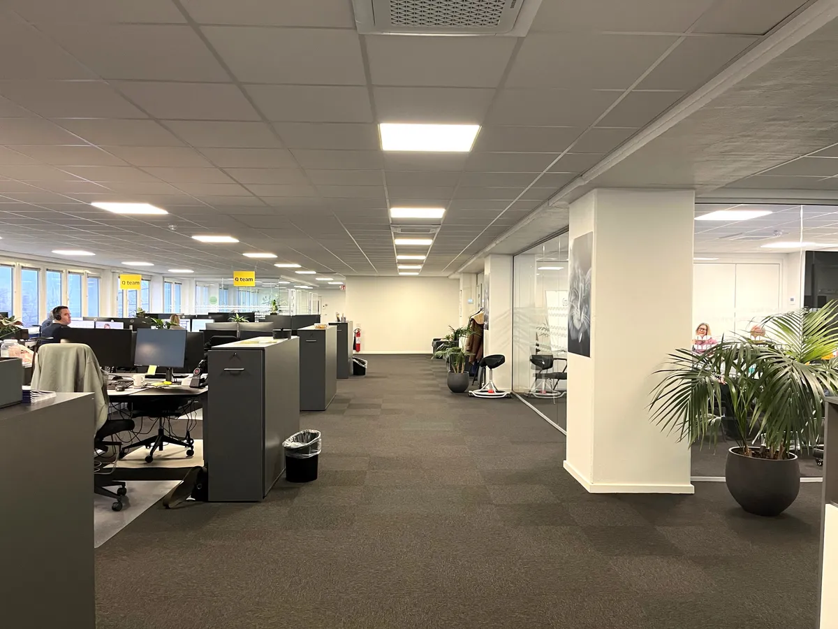 Kontor med glasvægge og møbler. Et enkelt menneske sidder på en stol. Et bord med computer og kuvert på. Planter og