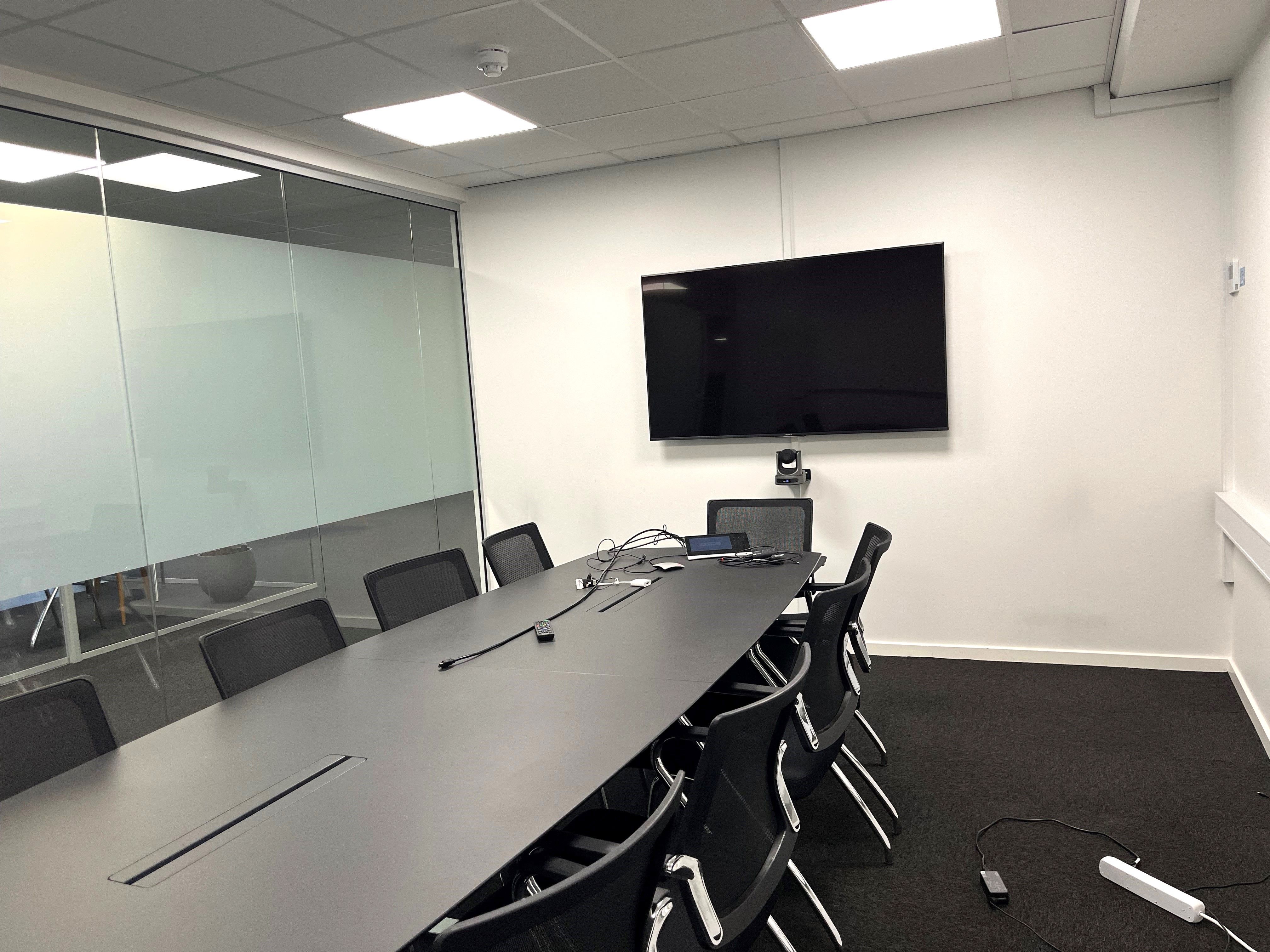 Kontorrum med lange borde og stole og en tv-skærm på væggen. Kabel og en lille monitor på bordet, og en lille hvid enhed med