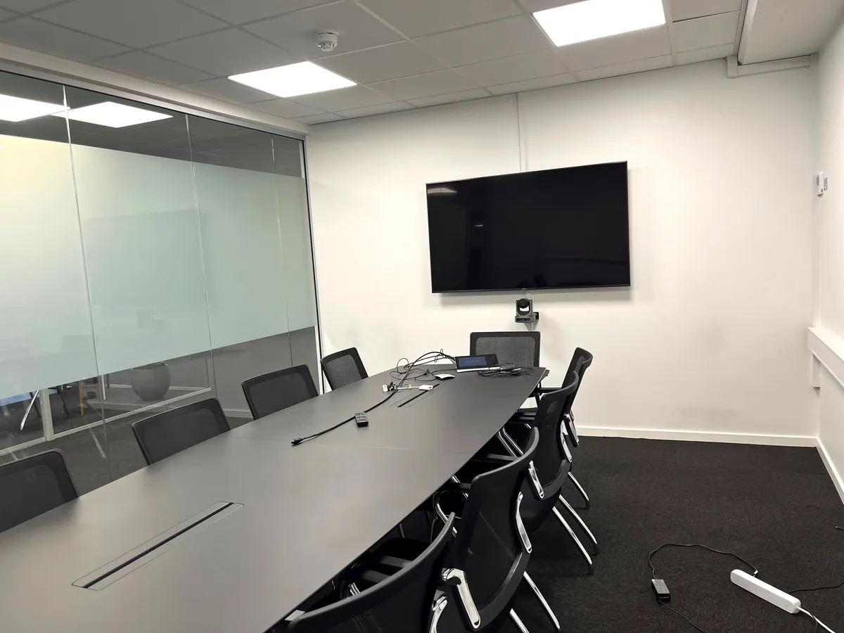 Kontorrum med lange borde og stole og en tv-skærm på væggen. Kabel og en lille monitor på bordet, og en lille hvid enhed med