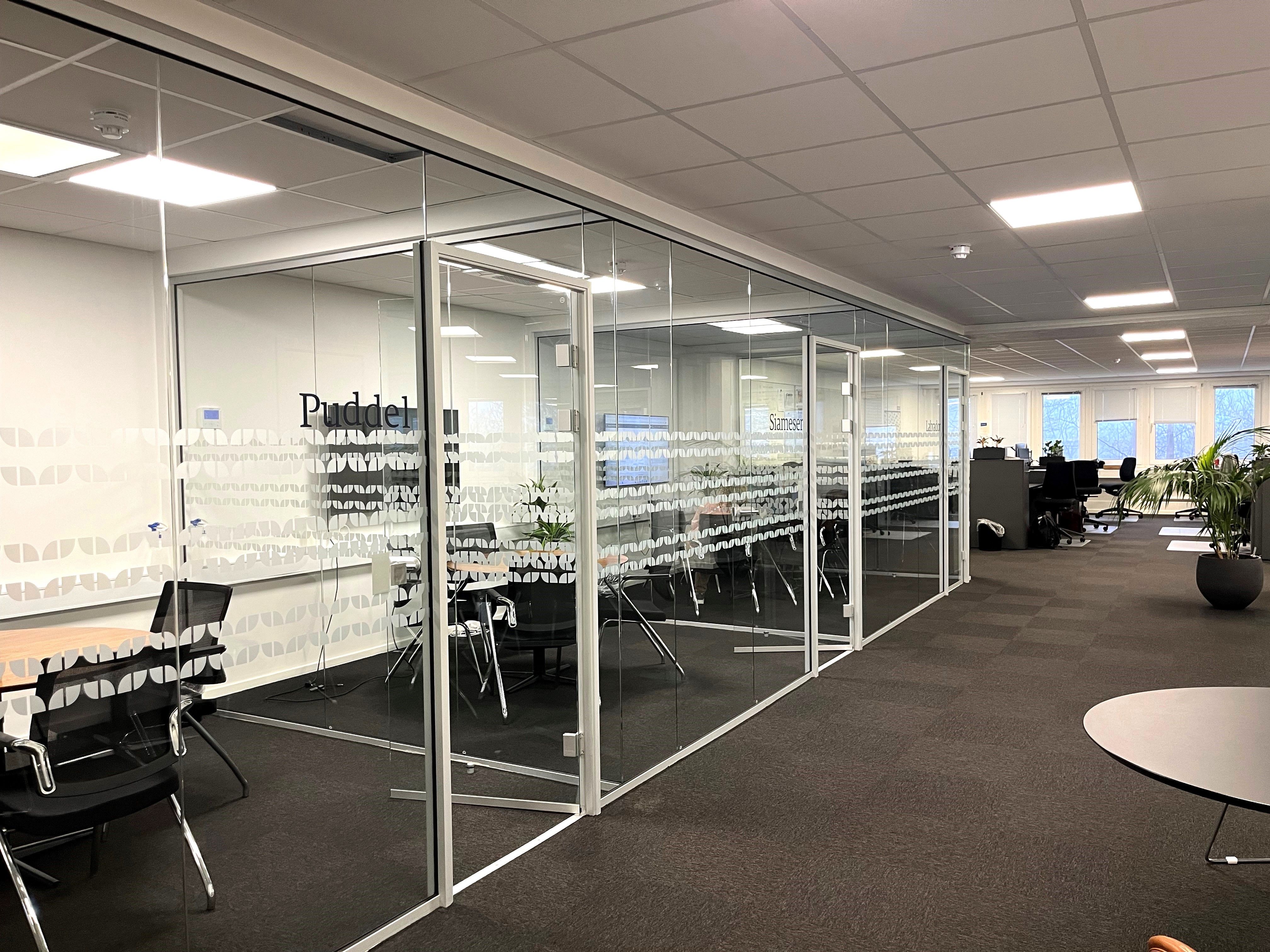 Kontor med glasvægge, hvide vægge, bord og stole, plante i gryde og lysekroner i loftet