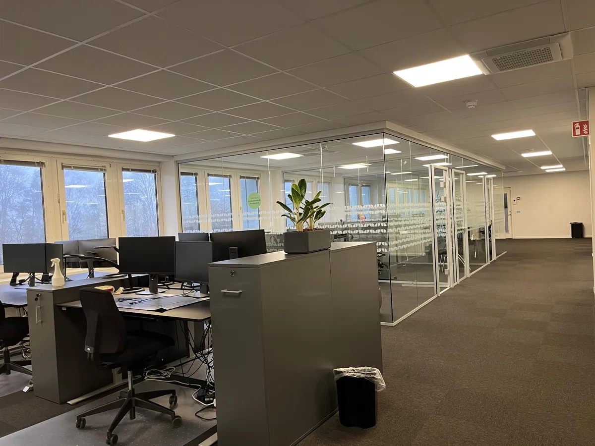 Kontorrum med lyse glasvægge og gulvbelægning samt flere computere og stole samt en plante i en potte