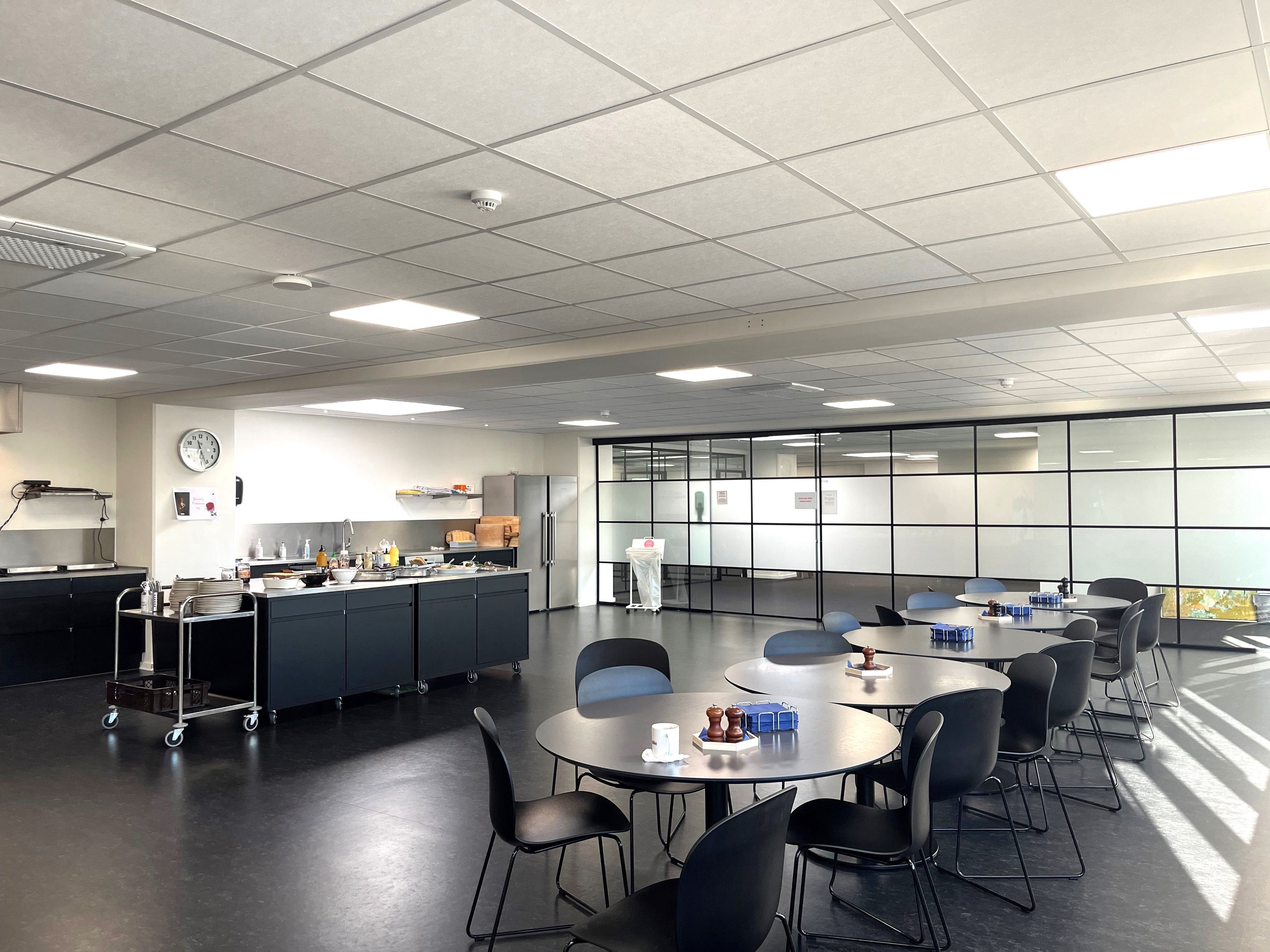 Kanteen er rummelig med møbler og borde. Der er en køkkenvæg med en række apparater og et ur på væggen