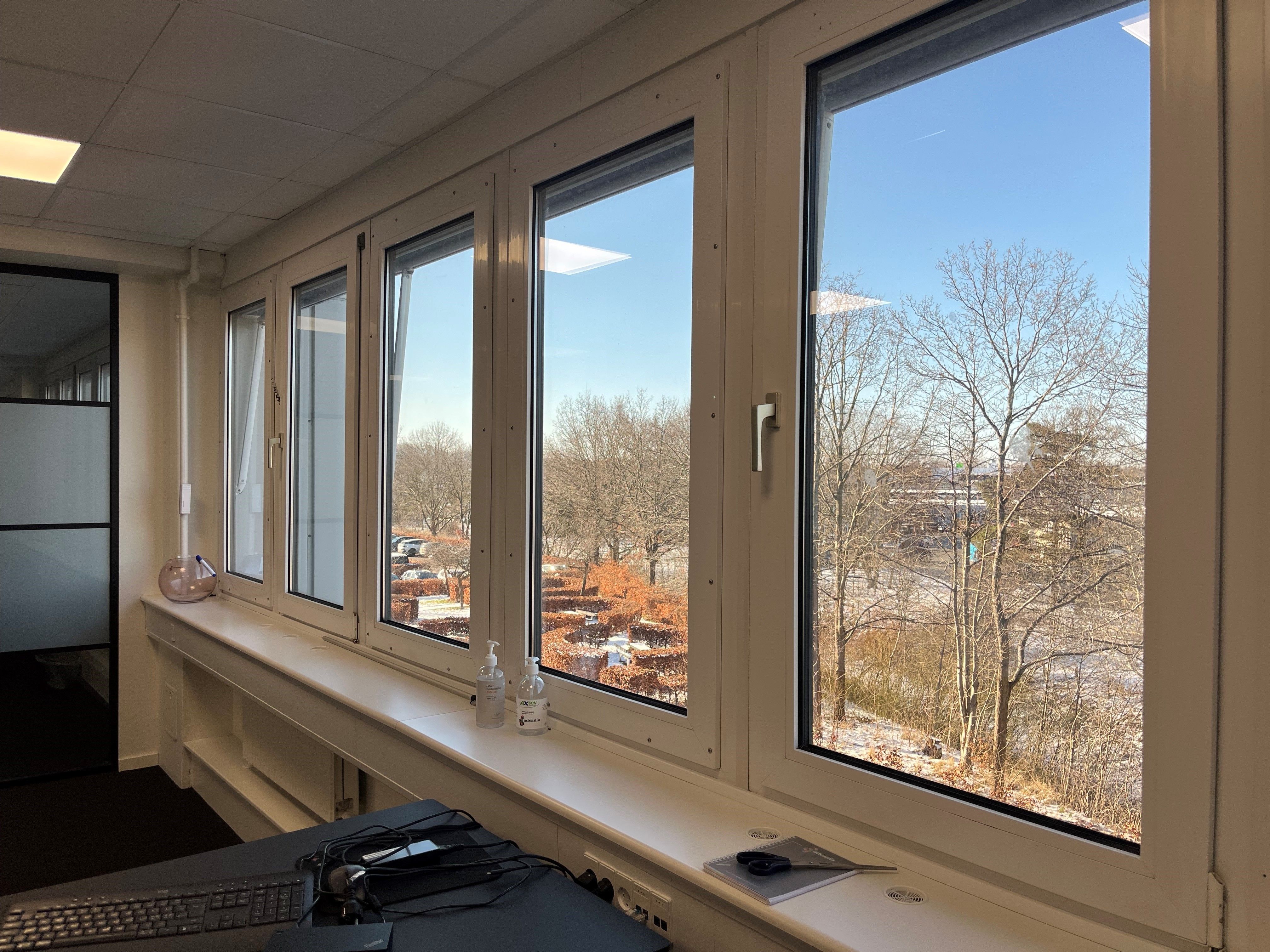 Glasvinduer med udsigt til et parkanlæg med bartrætræer, snedække og parkeringspladser i et moderne kontor med skrivebord,