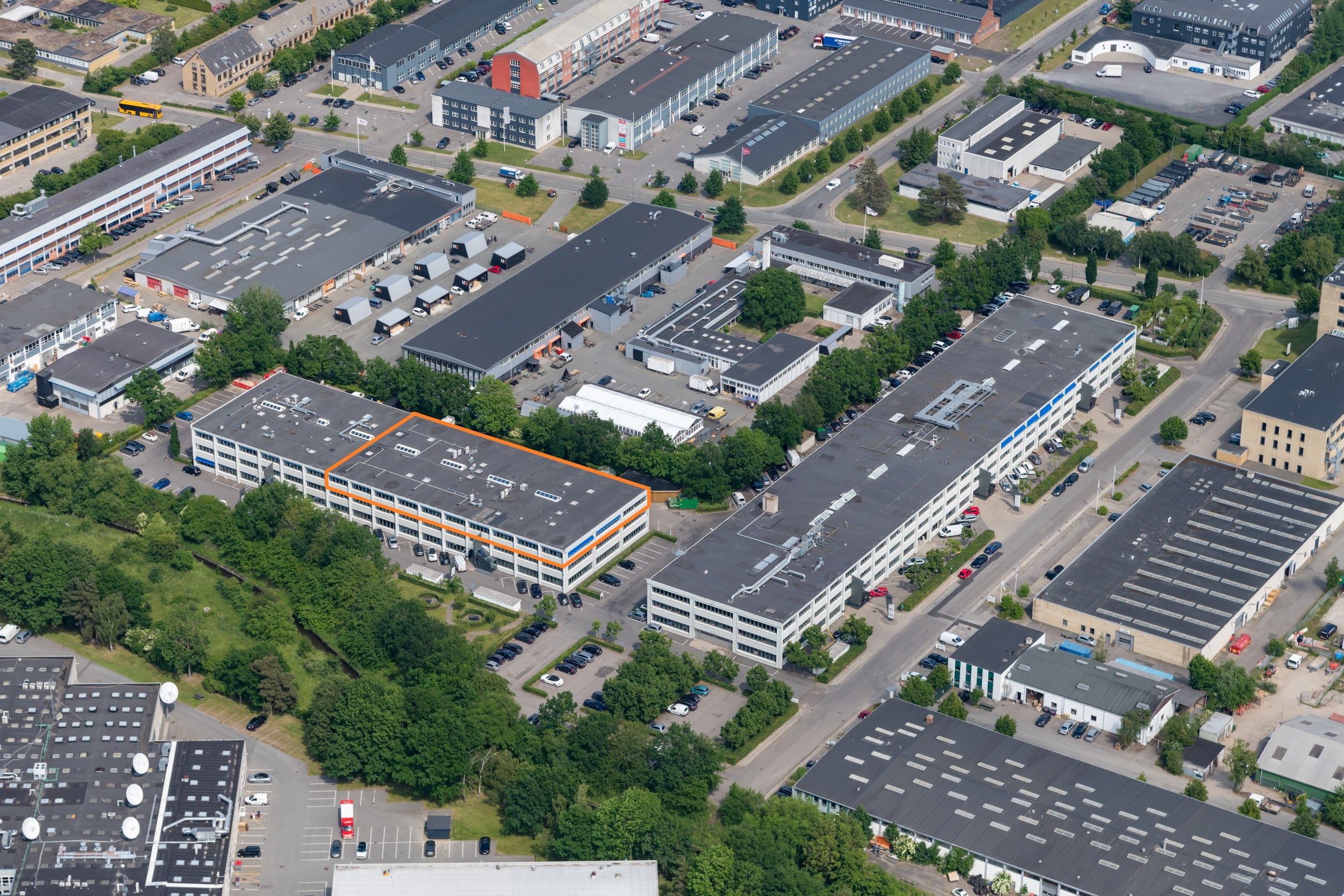 En stor bygning med orange kanterne og store glasruder, der er omgivet af grønne træer og andre bygninger