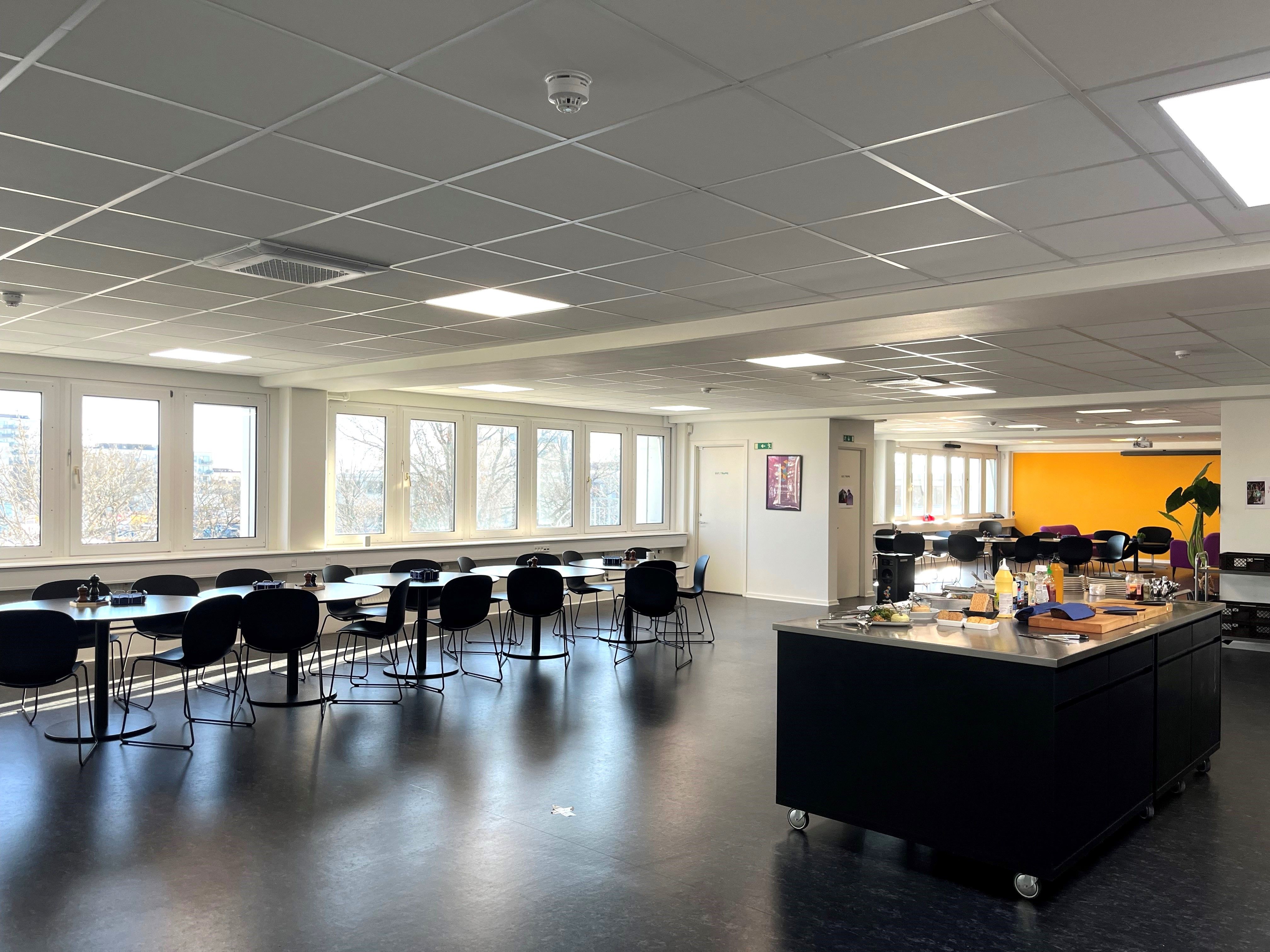 Kontorområde med lyse lofter, glasvinduer, bord og stol, samt et køkken med apparater og snitbræt