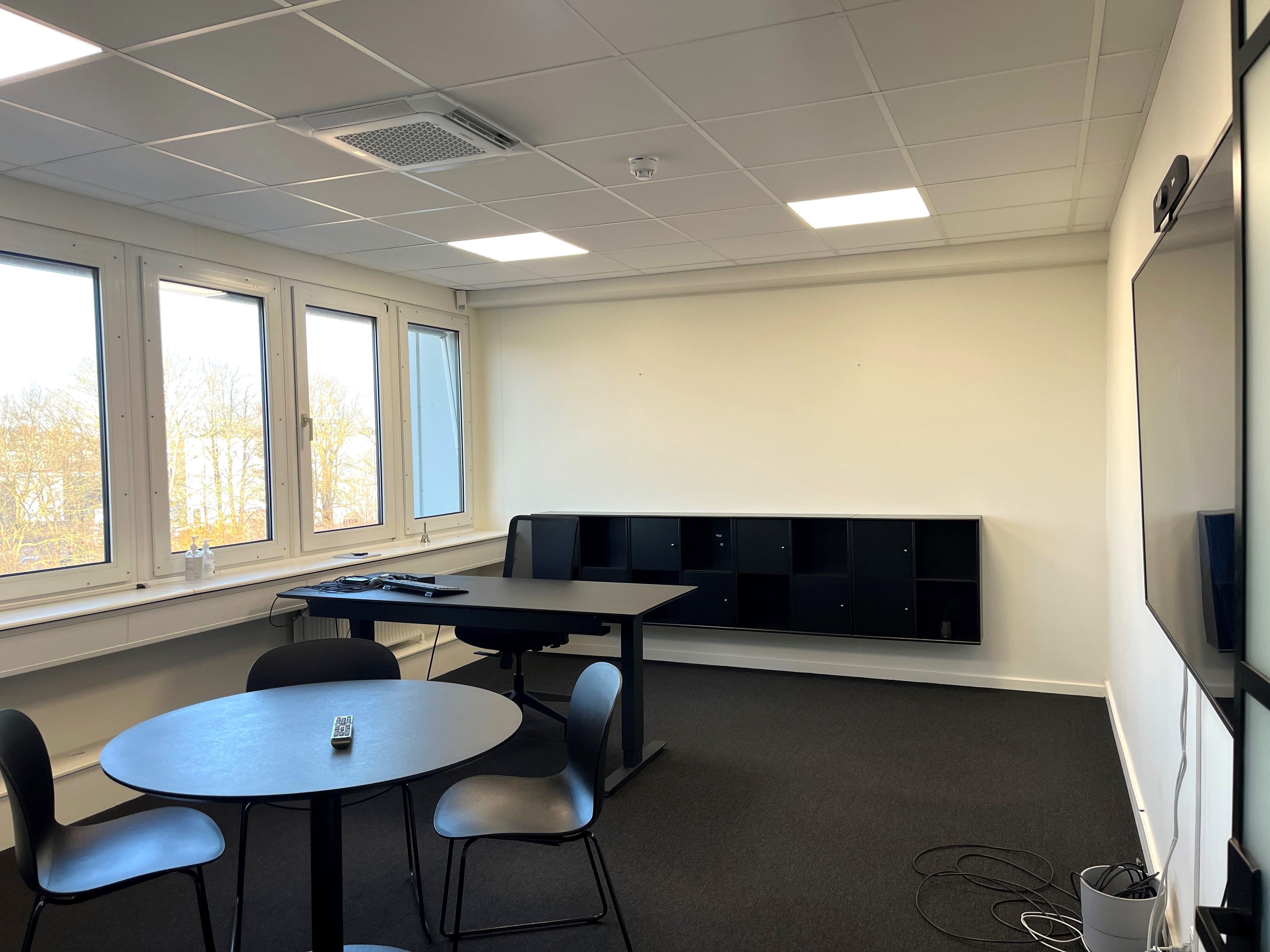 Kontorrum med store vinduer, mørke skriveborde, sorte stole, to rundere borde og tæt gulvbelægning