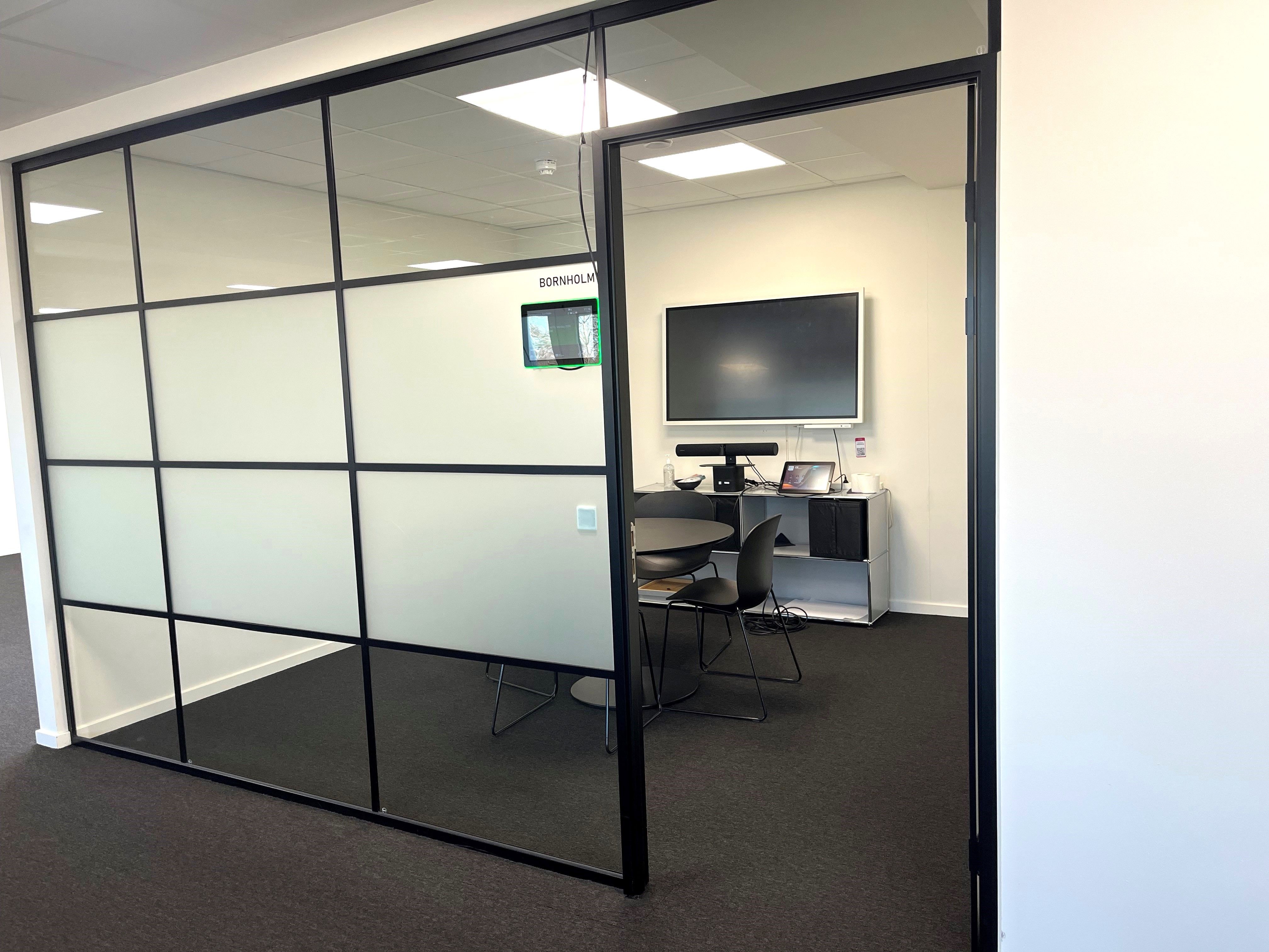 Kontorlokaler med glasvægge og møbelsæt, tv og skrivebord med en bærbar computer og flere stolpe.