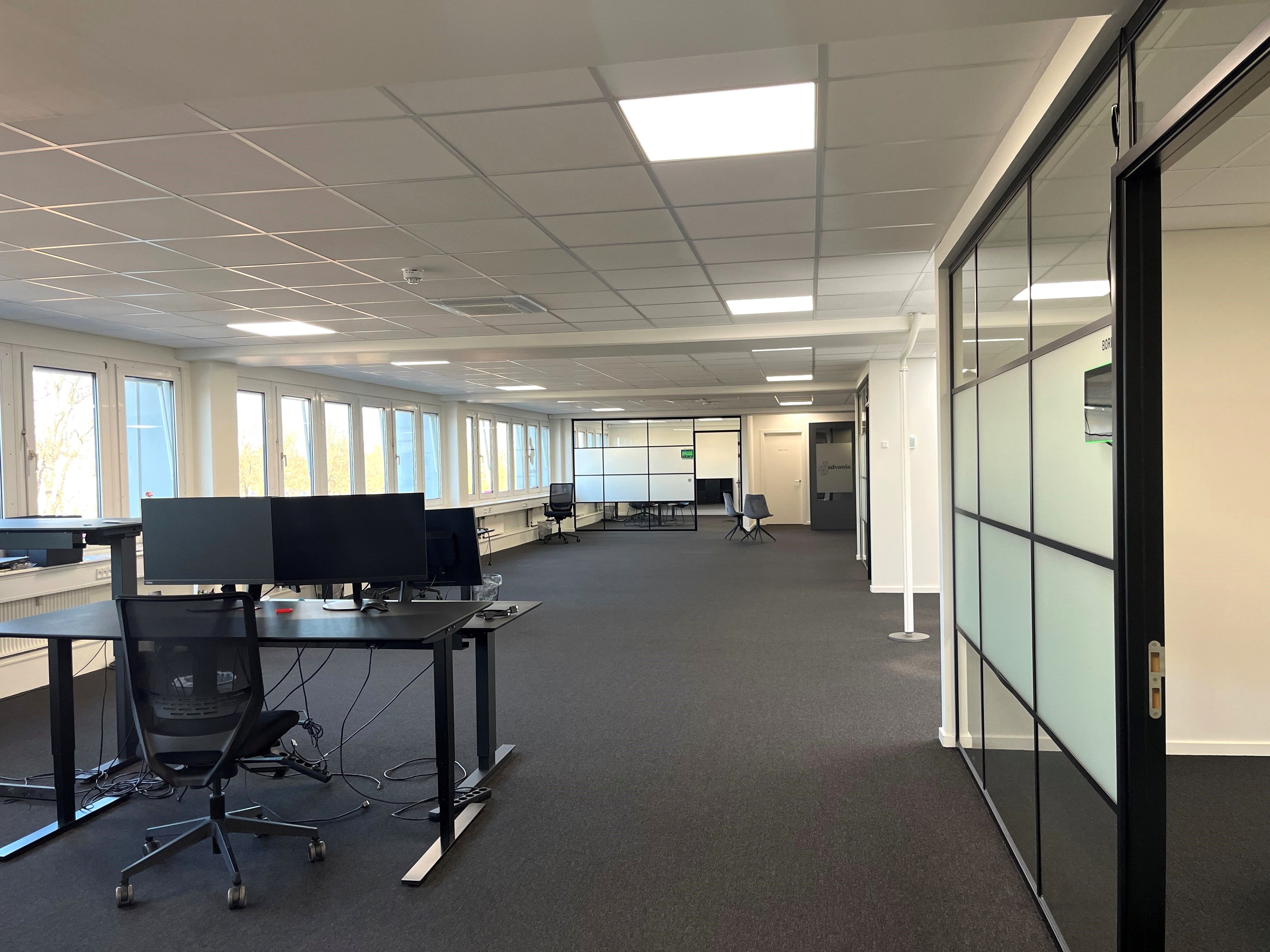 Kontor med skriveborde, skærme og lænestol i rum med glasvinduer og hvide vægge samt klimaanlæg