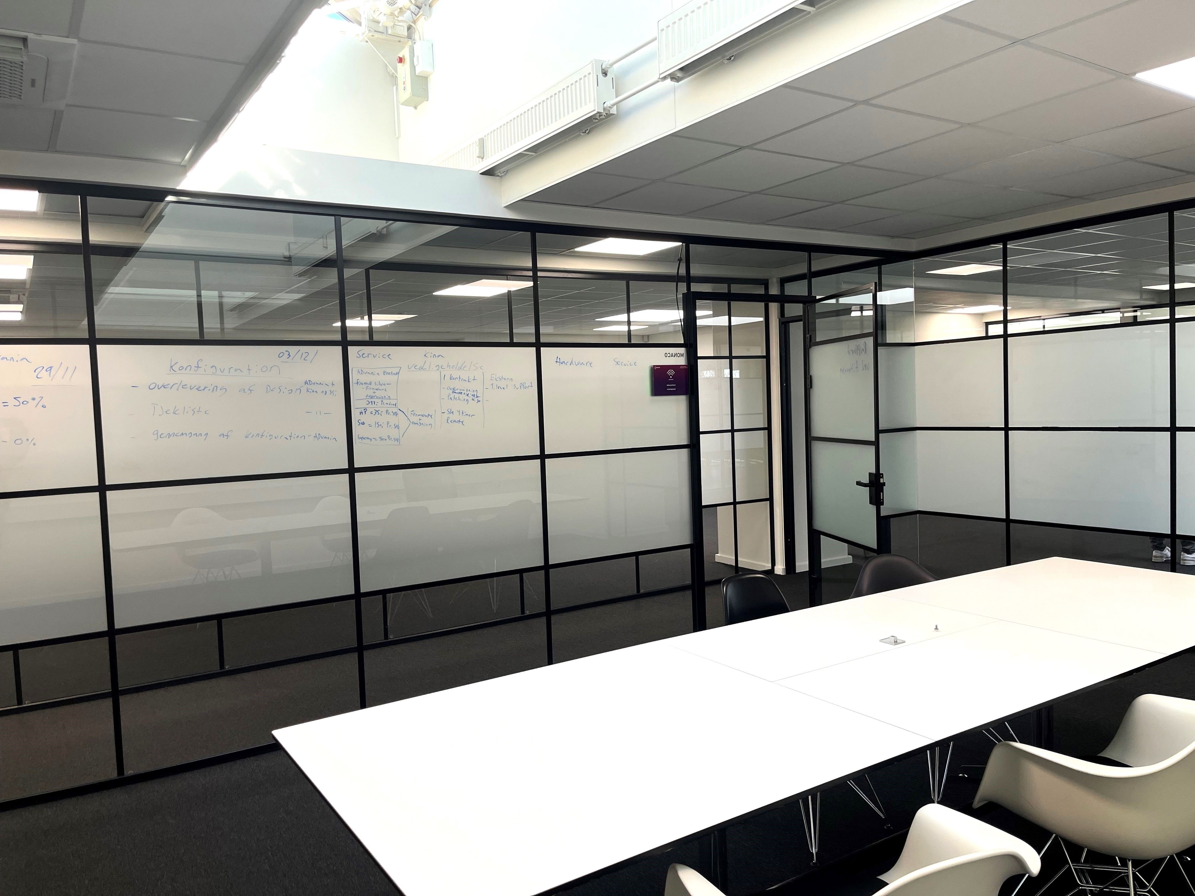Kontorlokaler med hvide borde og stolpeglasløsninger i sort. Væggen med whiteboards og flere noter. Foran er to hvide stole