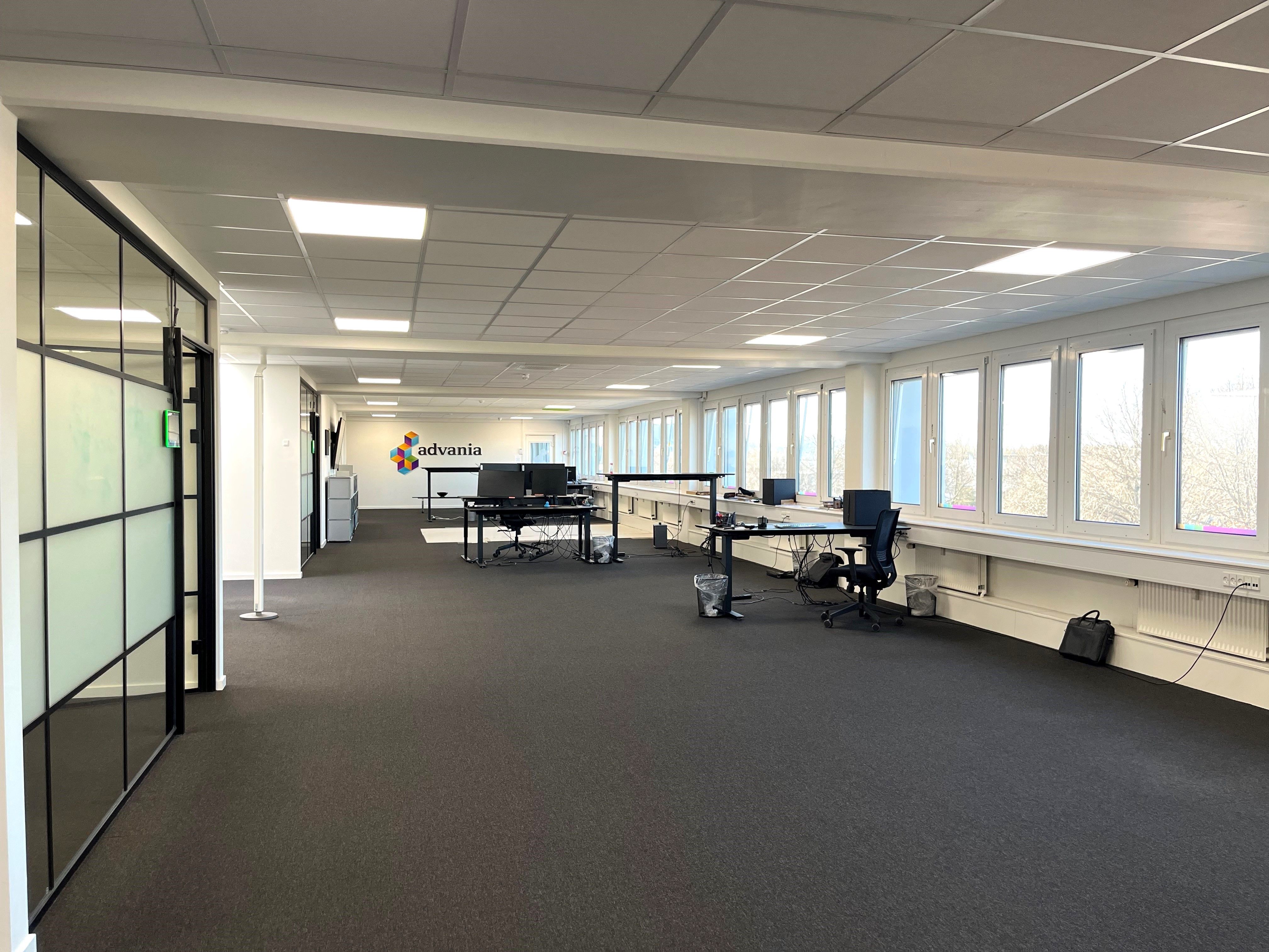 Moderne kontorlokaler med lyse vinduer og to skriveborde, hver med computer og skærm, samt en lille opbevaringsskranke i