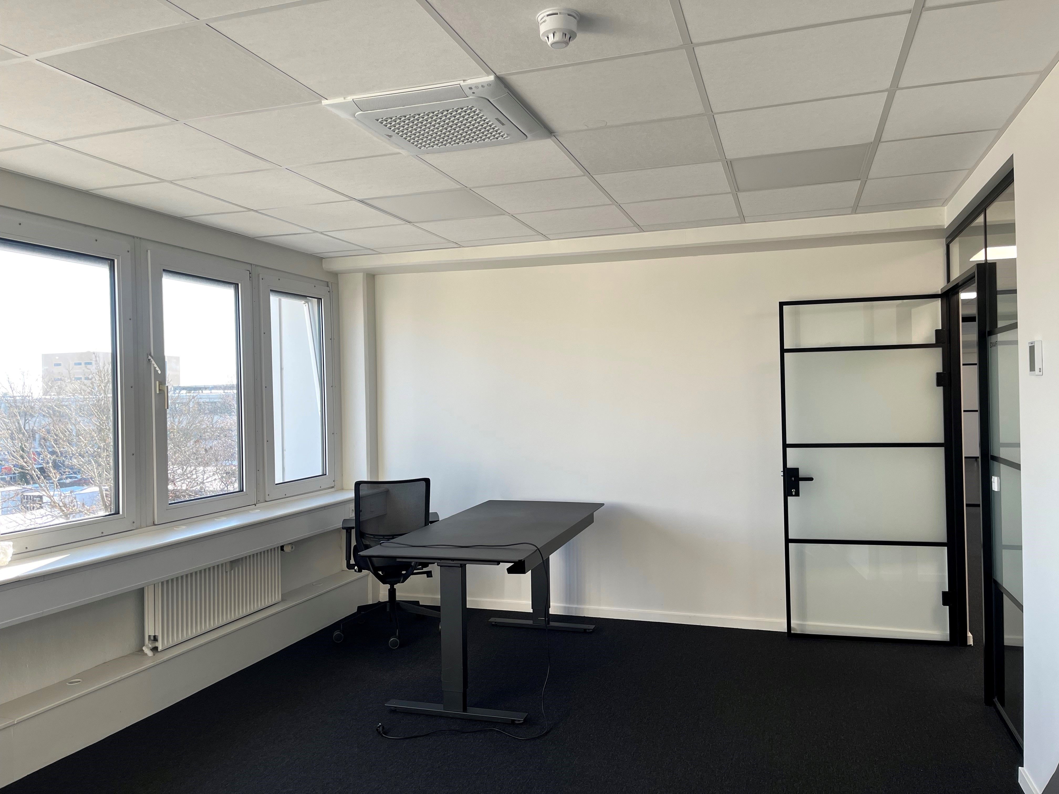 Kontorrum med skrivebord, stol, radiator, vinduer, klimaanlæg og glasdør. Kantineområde og lagerlokaler. Luftrensende