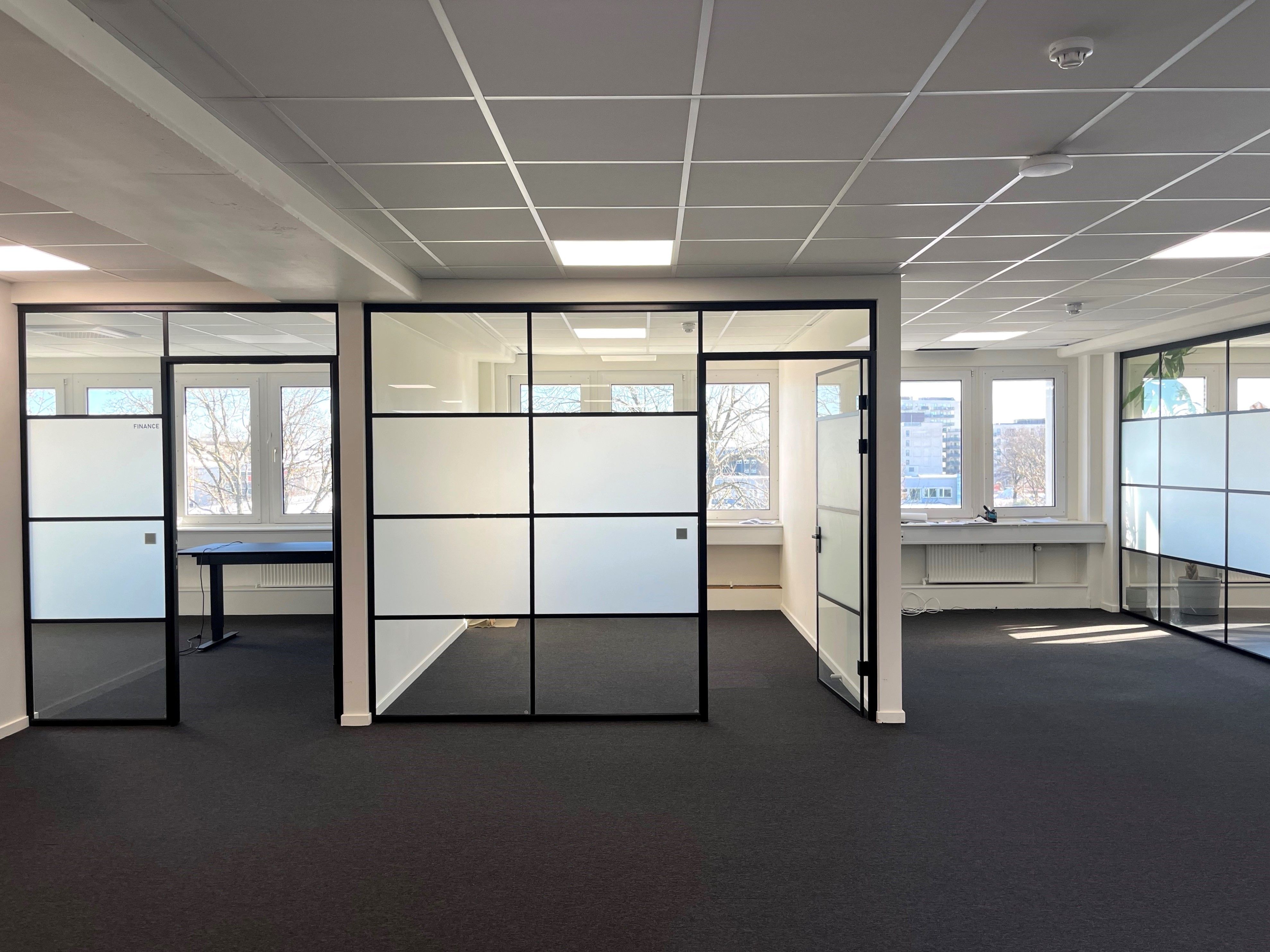 Kontor med glasvægge, gulvbelægning og klimaanlæg, to vinduer med udsigt og en lille sofa.