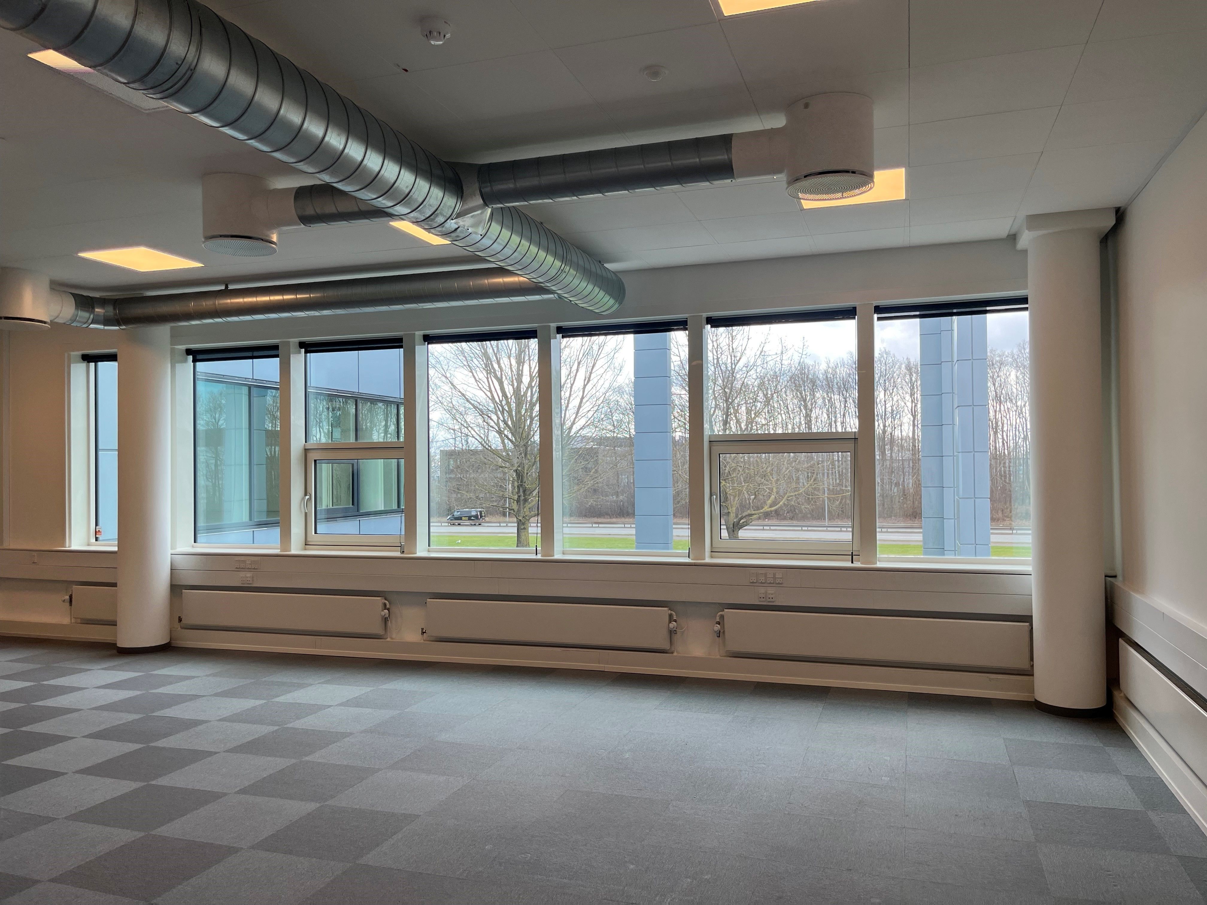 Et stort kontor med vinduer, hvor der står flere radiatorer og store rør i loftet. Gulvet er lavet af grå og hvide krydsfiner