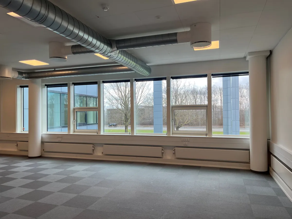 Et stort kontor med vinduer, hvor der står flere radiatorer og store rør i loftet. Gulvet er lavet af grå og hvide krydsfiner