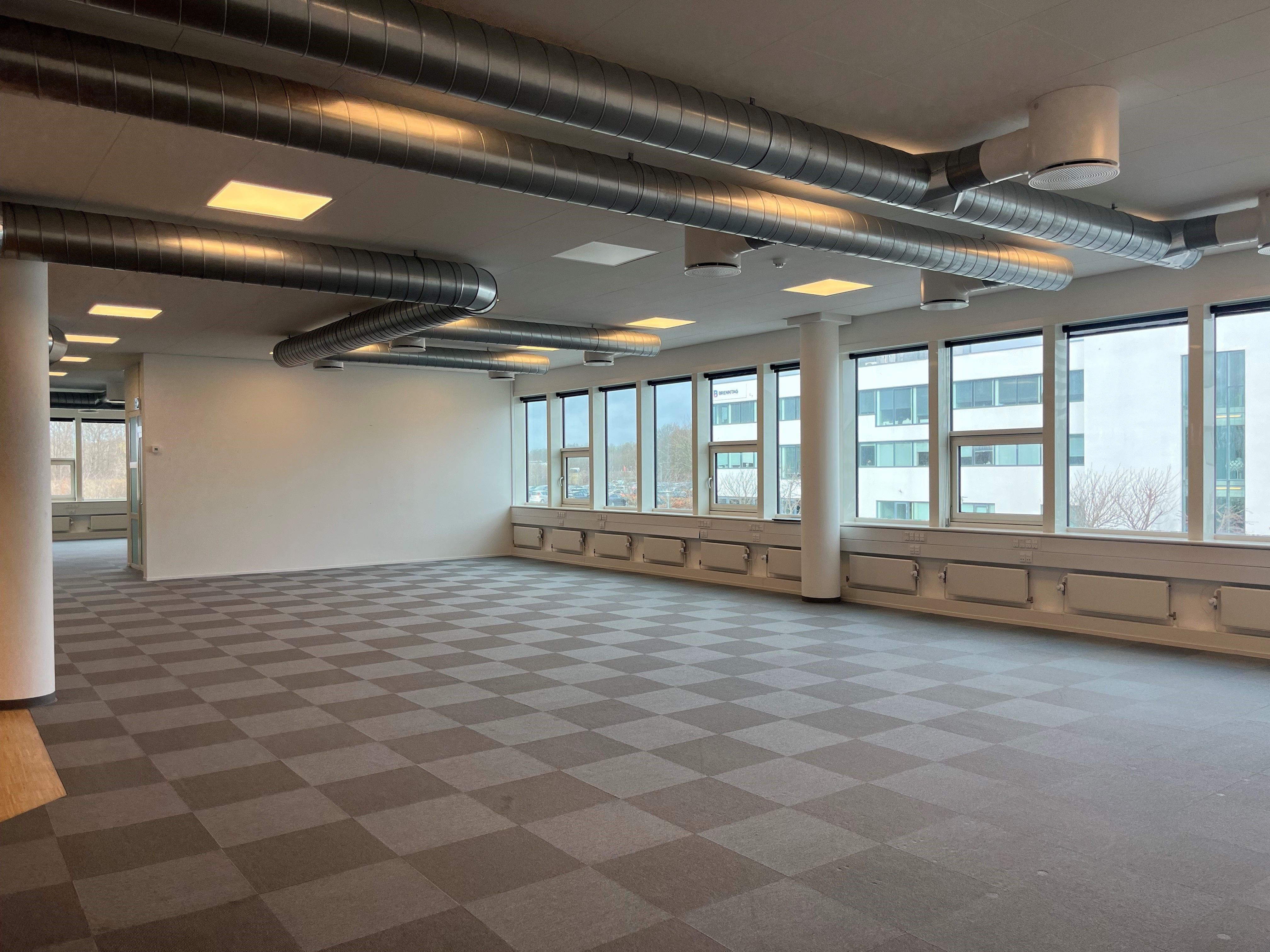 Foto af et stort og rummeligt kontor med karrevinduer, store rør i loftet, gulvbelægning i grønne og grå ruter og lyse