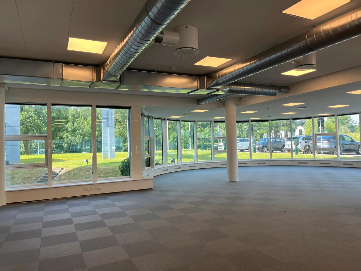 Kontor med glasvinduer og udsigt til park. Indvendigt med lysegult lys og slanke støttepiller. Parkerede biler og grønne