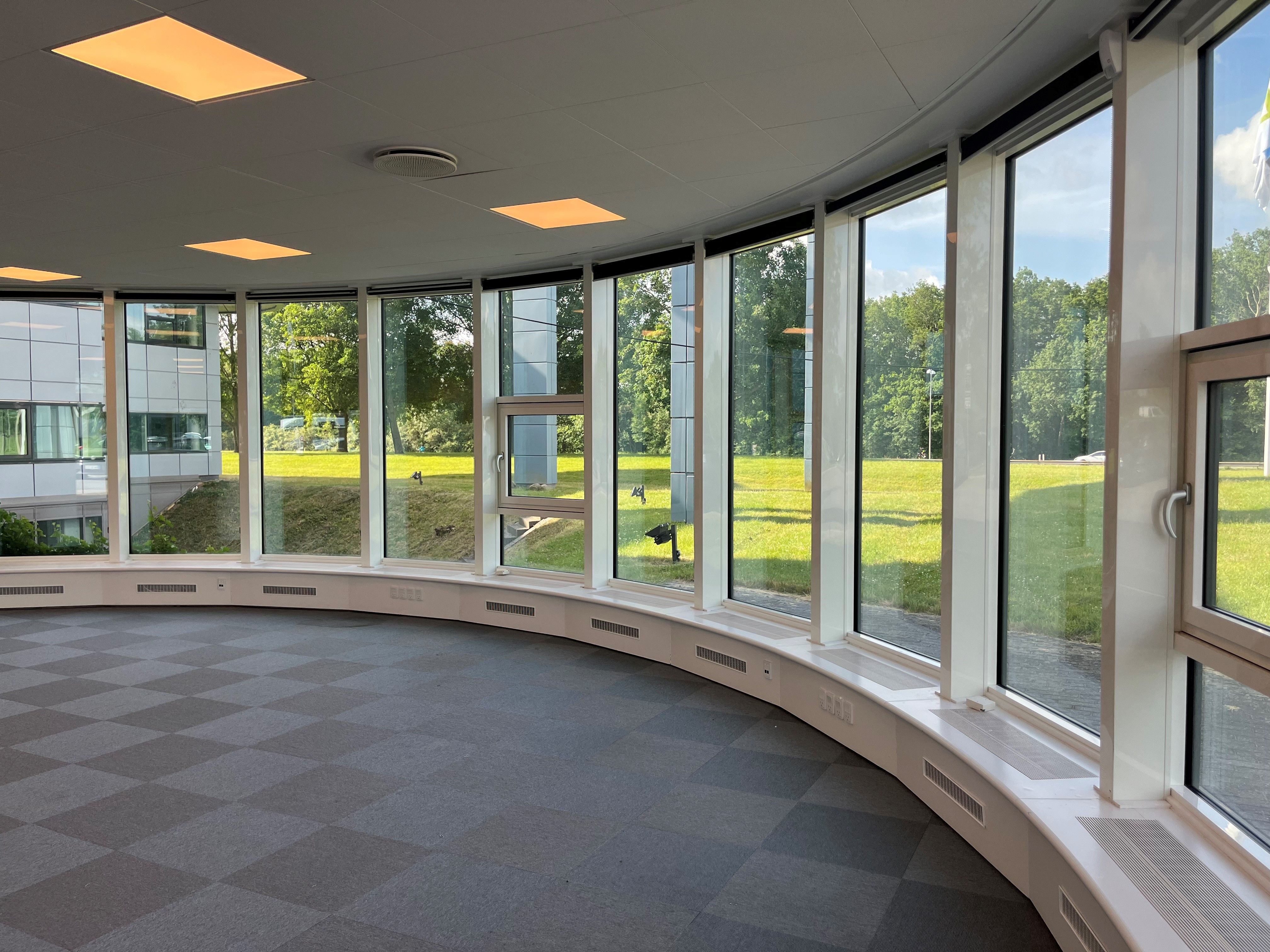 Kontor med store vinduer, runde gulv mønstre, hvide vægge, grønne områder udenfor og flere bygninger i baggrunden